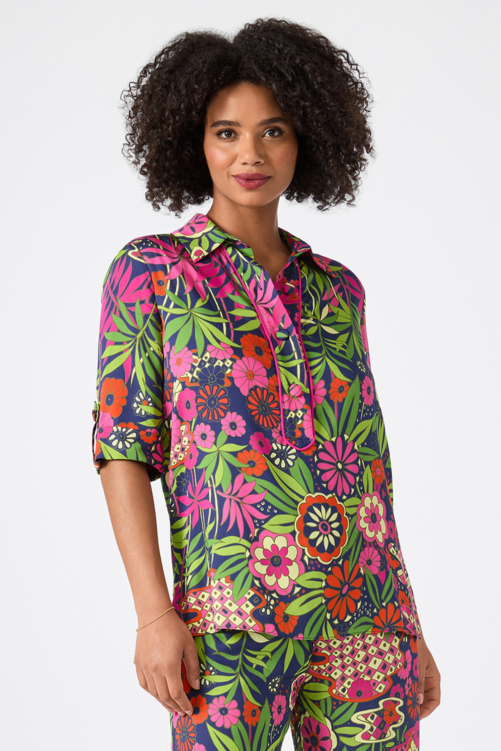 A woman with curly hair and a slight smile wears the Portrait 2 Top in Fumi Floral, featuring bold pink, red, green, and yellow flowers with leafy designs on a dark background.