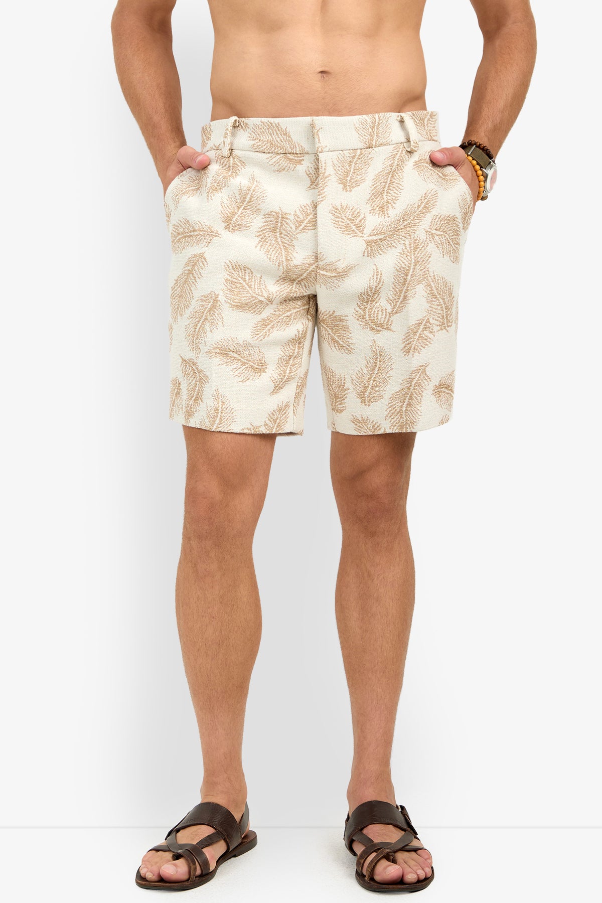 Someone wearing the Lawrence Short in beige with a brown leaf pattern, paired with brown sandals and a bracelet, stands against a white background with hands in their pockets.