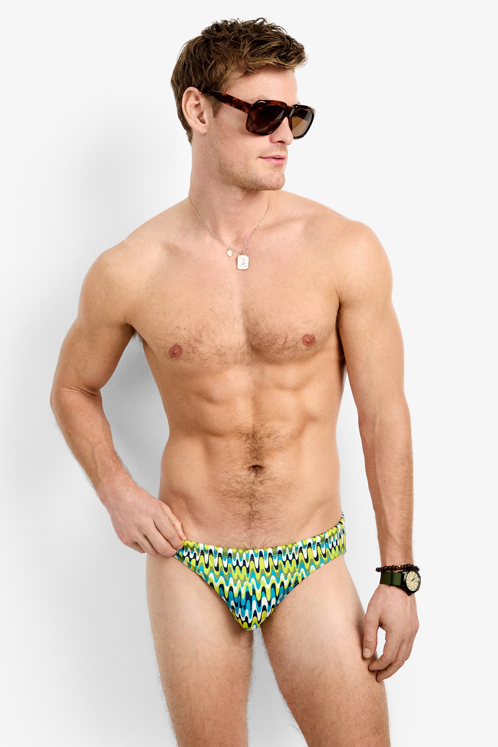 A man wearing sunglasses, a necklace, a wristwatch, and the Lagos Swim Brief - Psychedelic Voyage stands against a plain white background, looking off to the side.