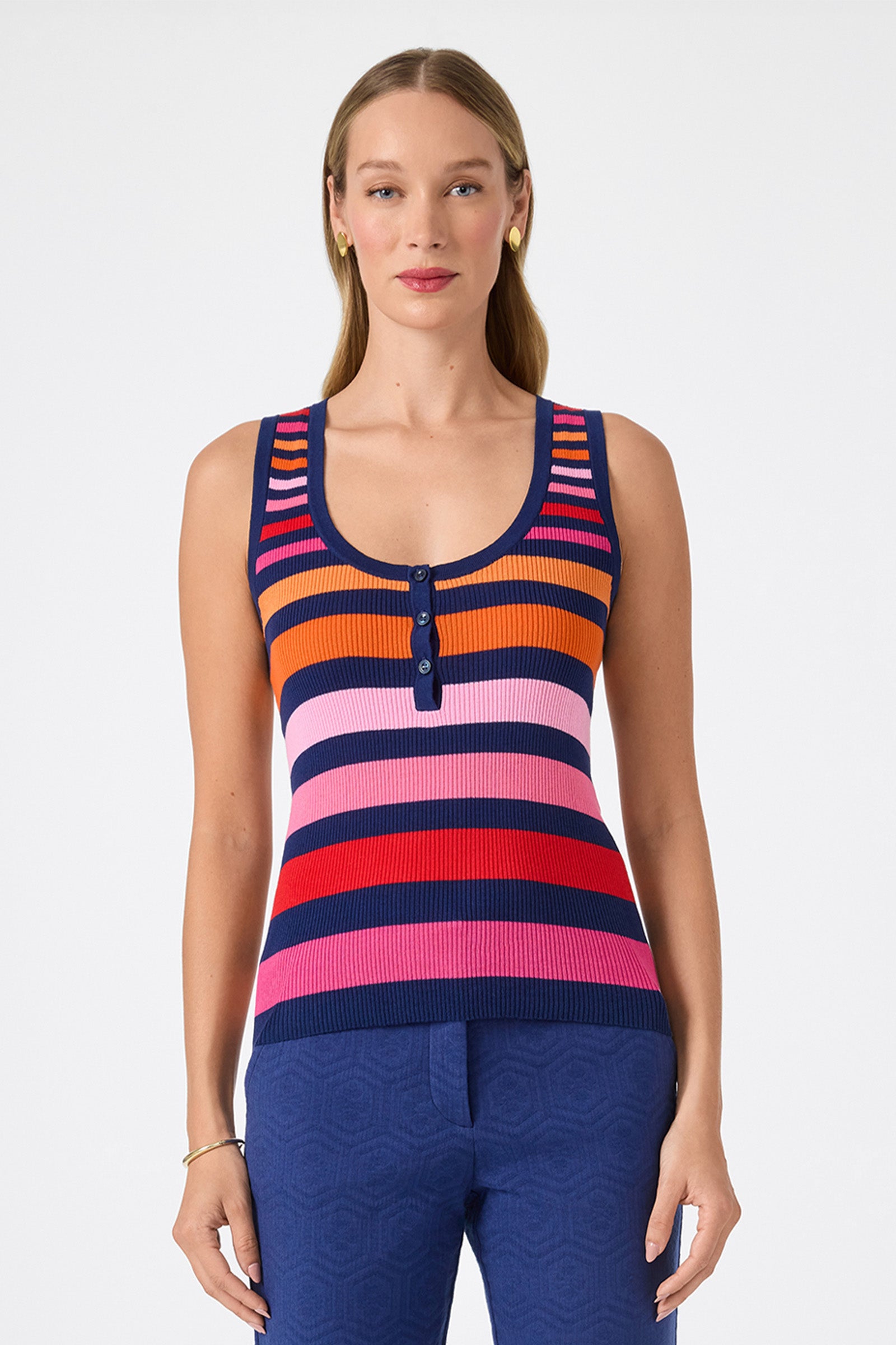A woman with long, straight blonde hair wears the Arles Scoop Neck Tank, a sleeveless multicolored striped knit top with buttons, paired with blue pants. She stands facing forward against a plain white background.