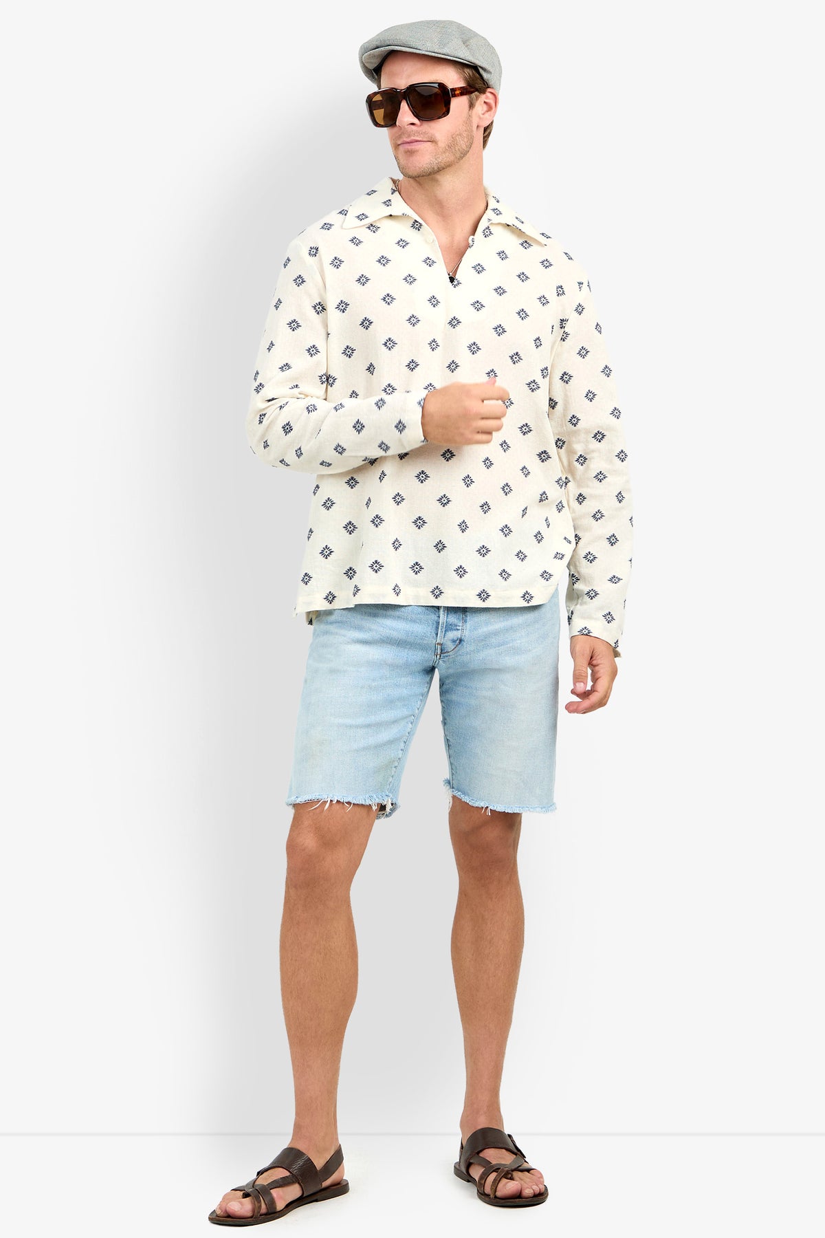 Man wearing sunglasses, a gray flat cap, Hendrix Long-Sleeve Pullover, light denim shorts, and brown sandals stands against a plain white background.
