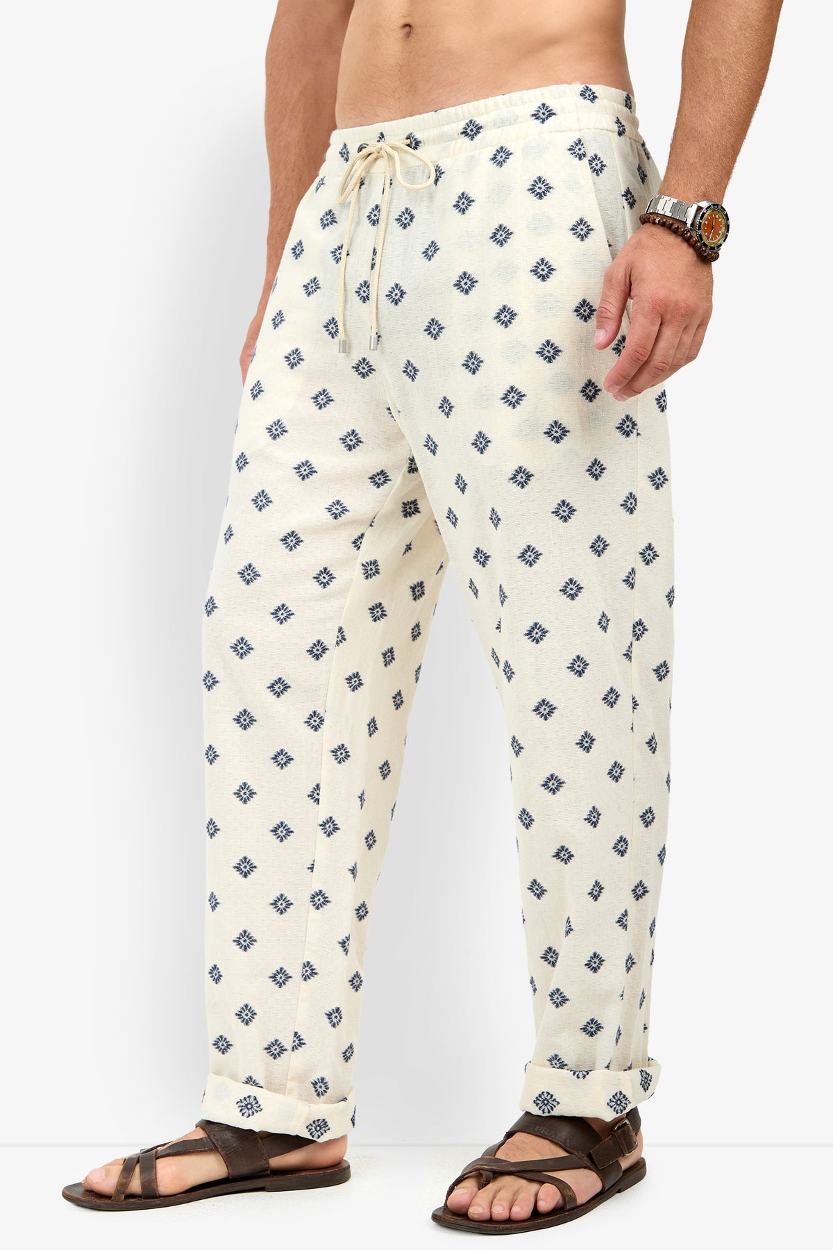 A person wearing the Felipe Drawstring Pant in cream with a blue geometric pattern, paired with brown sandals and a beaded bracelet, stands against a plain background.