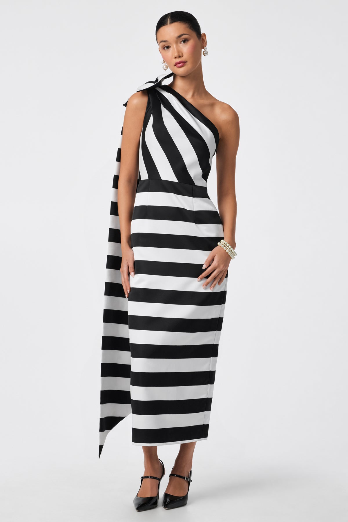 A woman models the Romance Dress, a black and white striped one-shoulder ankle-length gown with an asymmetrical drape, paired with black heels and pearl accessories, against a plain white background.
