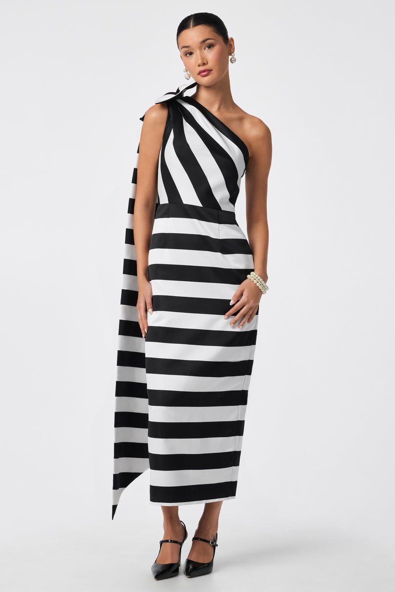 A woman models the Romance Dress, a black and white striped one-shoulder ankle-length gown with an asymmetrical drape, paired with black heels and pearl accessories, against a plain white background.