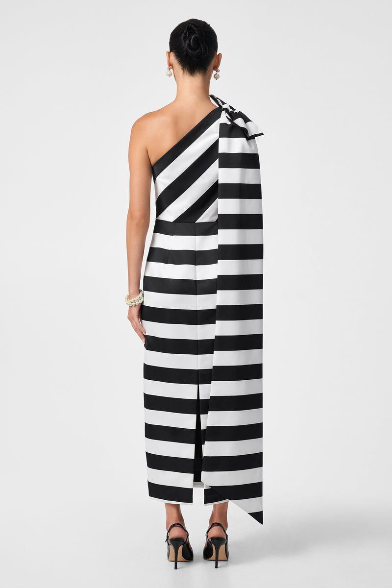 A woman, hair up, models the Romance Dress—a black and white horizontal striped one-shoulder style with a long draped fabric detail. She pairs it with black heels and a bracelet, posing against a plain backdrop.