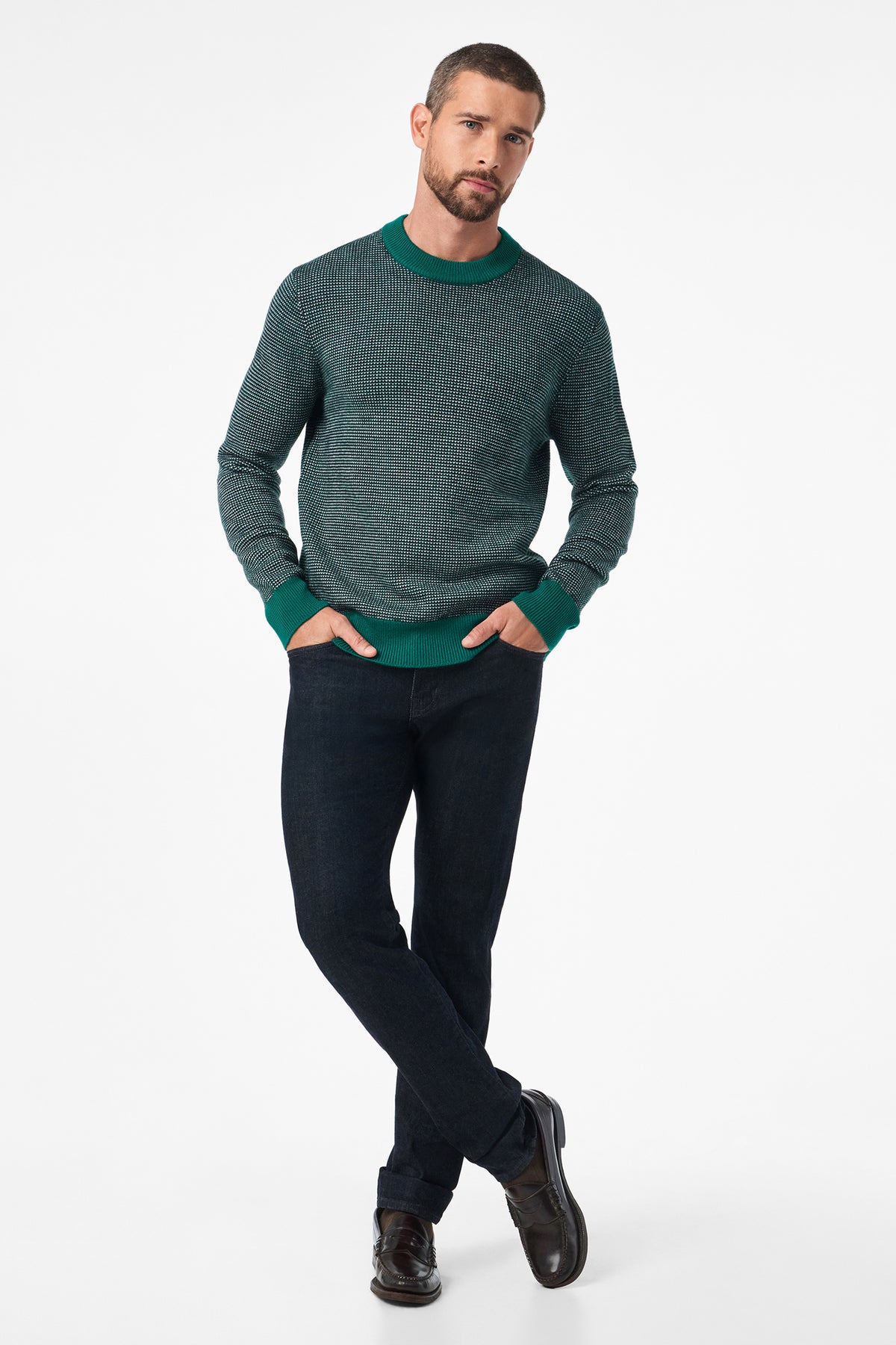 A man with short hair and a trimmed beard poses with his hands in his pockets, wearing the Ezra Crewneck in green, dark jeans, and dark shoes against a plain white background.
