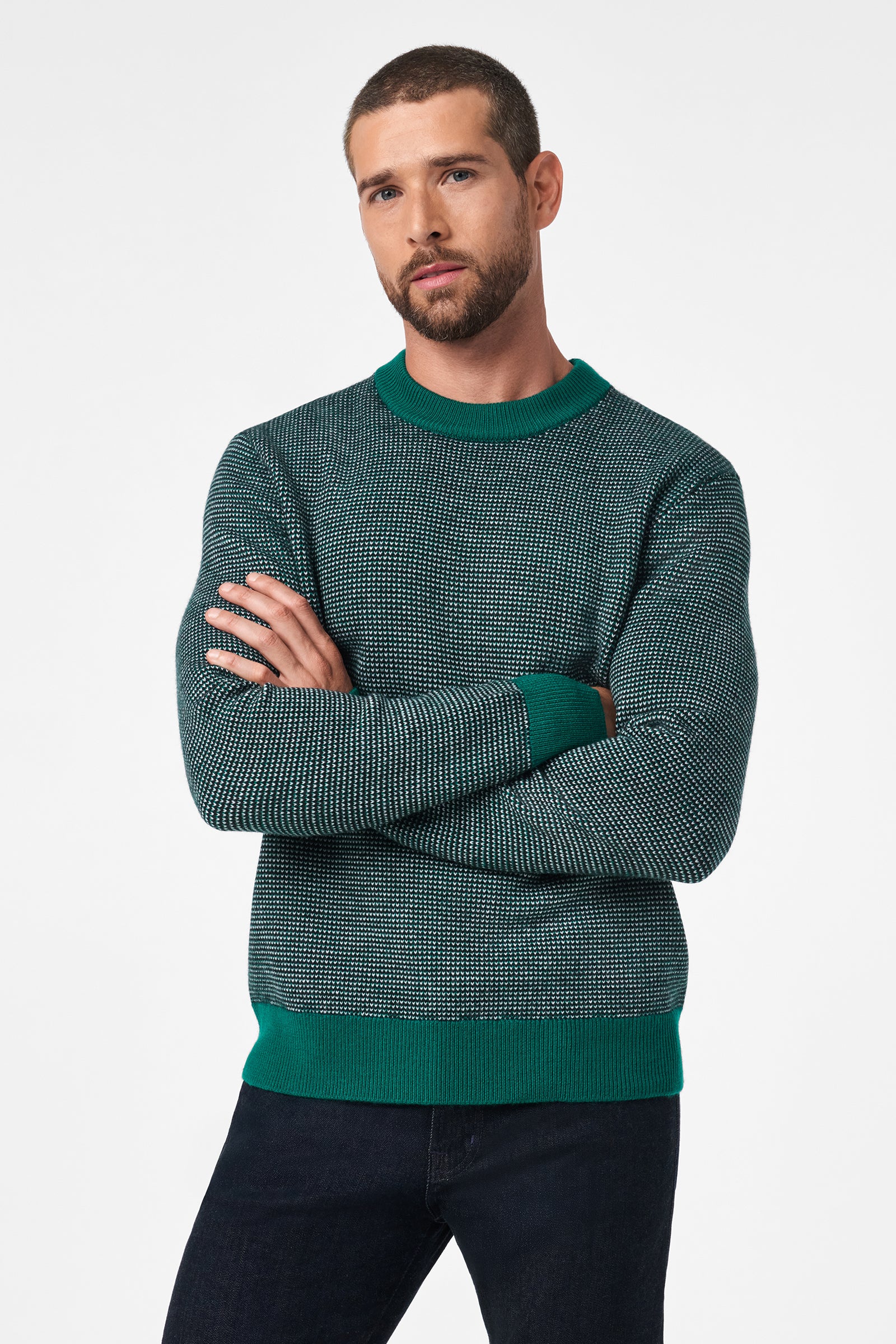 A man with short hair and a beard stands with his arms crossed, wearing the Ezra Crewneck—a green and black patterned sweater with matching green cuffs, collar, and hem—paired with dark jeans against a plain white background.