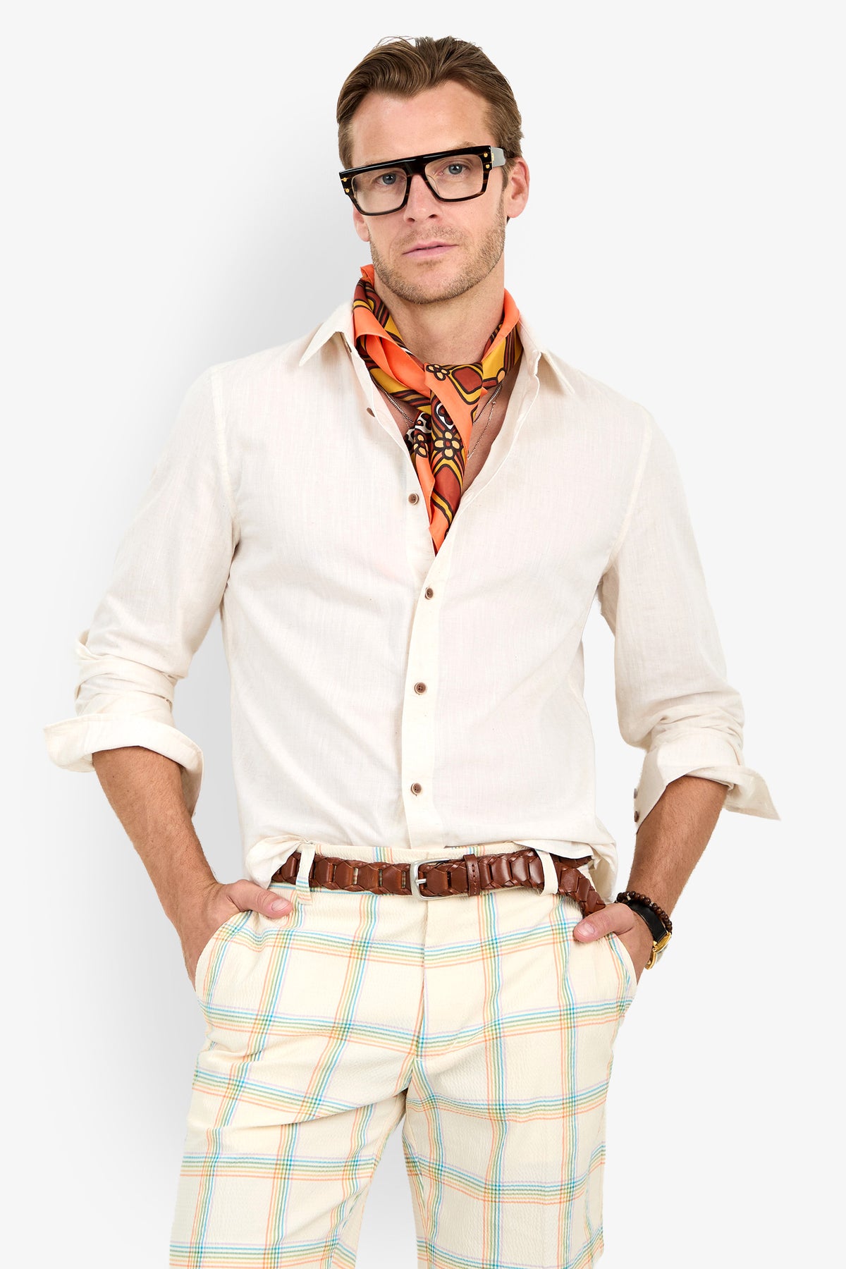 A man wears the Aaron Shirt in cream, paired with plaid pants, dark glasses, and an orange patterned scarf, standing with hands in pockets against a plain white background.