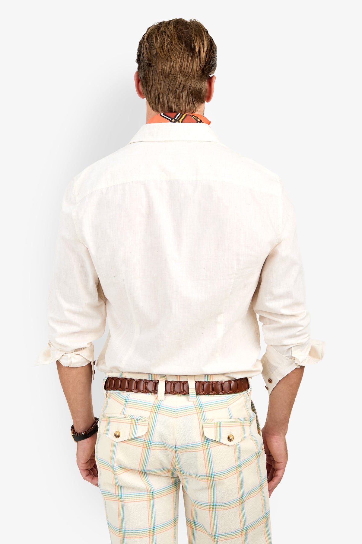 A man stands facing away, wearing the Aaron Shirt with rolled-up sleeves, plaid pastel-striped pants, a brown belt, bracelet, and a colorful scarf around his neck.