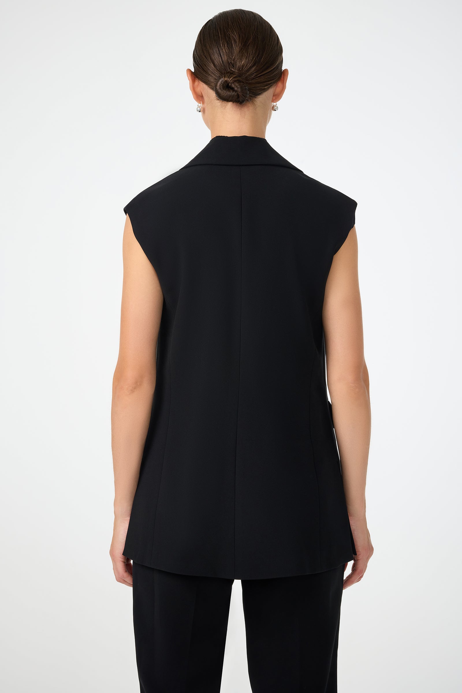 A person with neatly styled hair in a low bun is shown from behind, wearing the Boabab Sleeveless Jacket and black trousers against a plain light background.