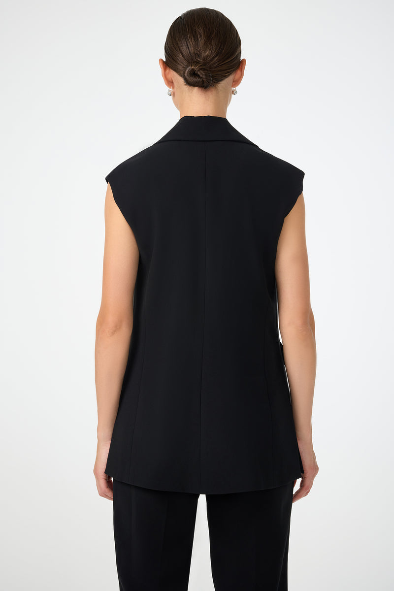 A person with neatly styled hair in a low bun is shown from behind, wearing the Boabab Sleeveless Jacket and black trousers against a plain light background.
