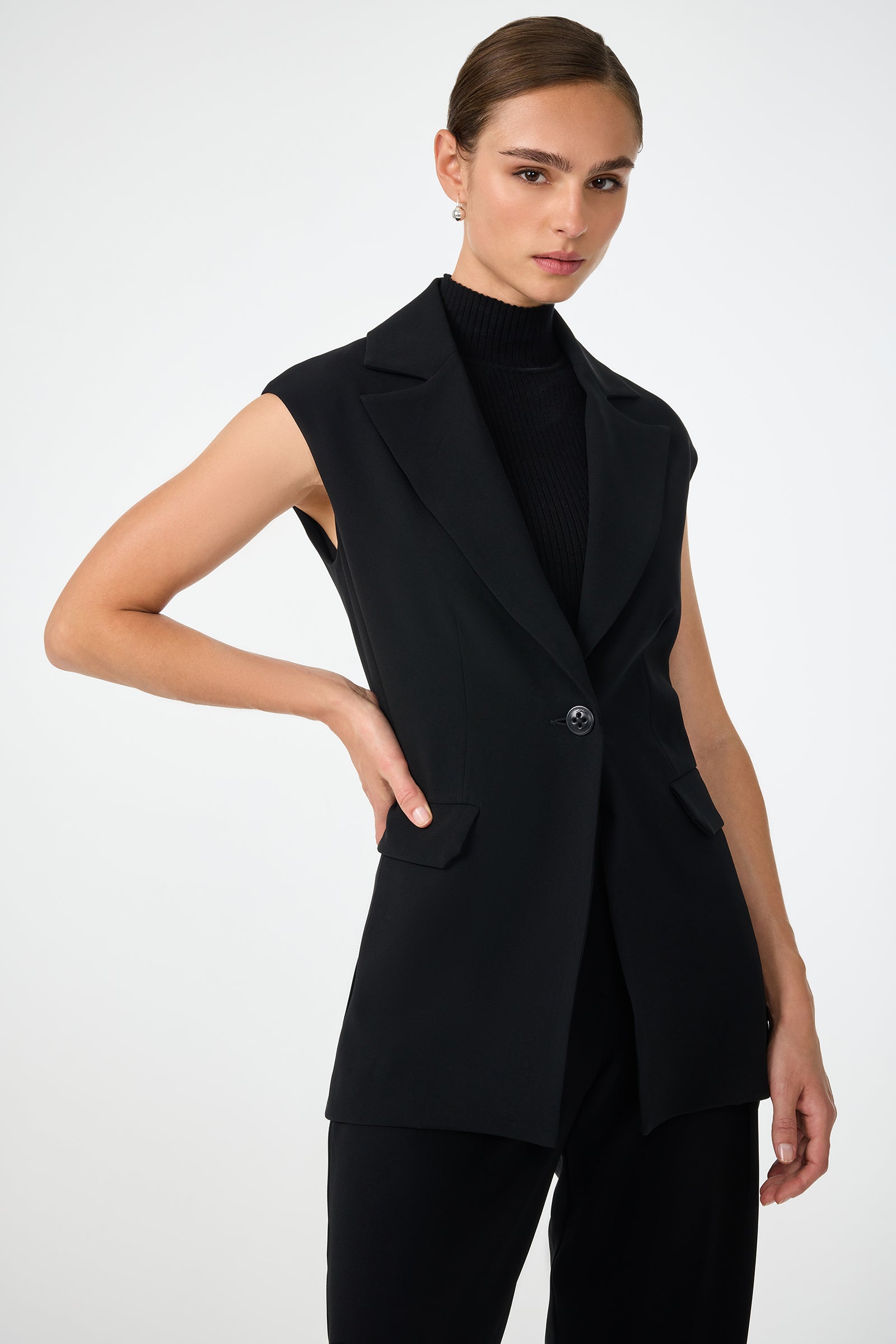 A woman with brown hair in a sleek bun wears the black Boabab Sleeveless Jacket over a black turtleneck and pants, standing with one hand on her hip against a white background.