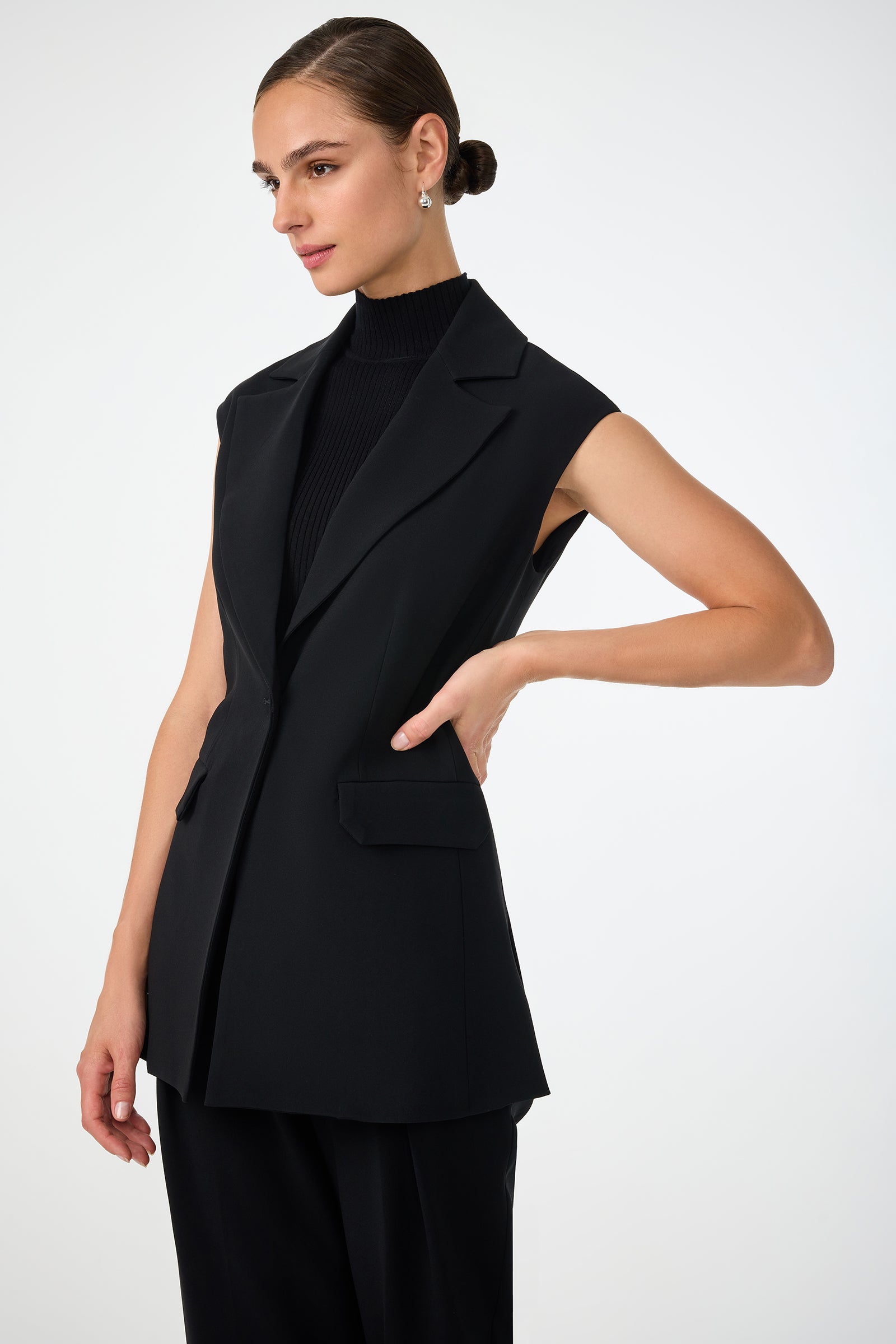A woman with dark hair in a low bun wears the Boabab Sleeveless Jacket over a black turtleneck and pants. She poses with one hand on her waist and looks to the side against a plain light background.