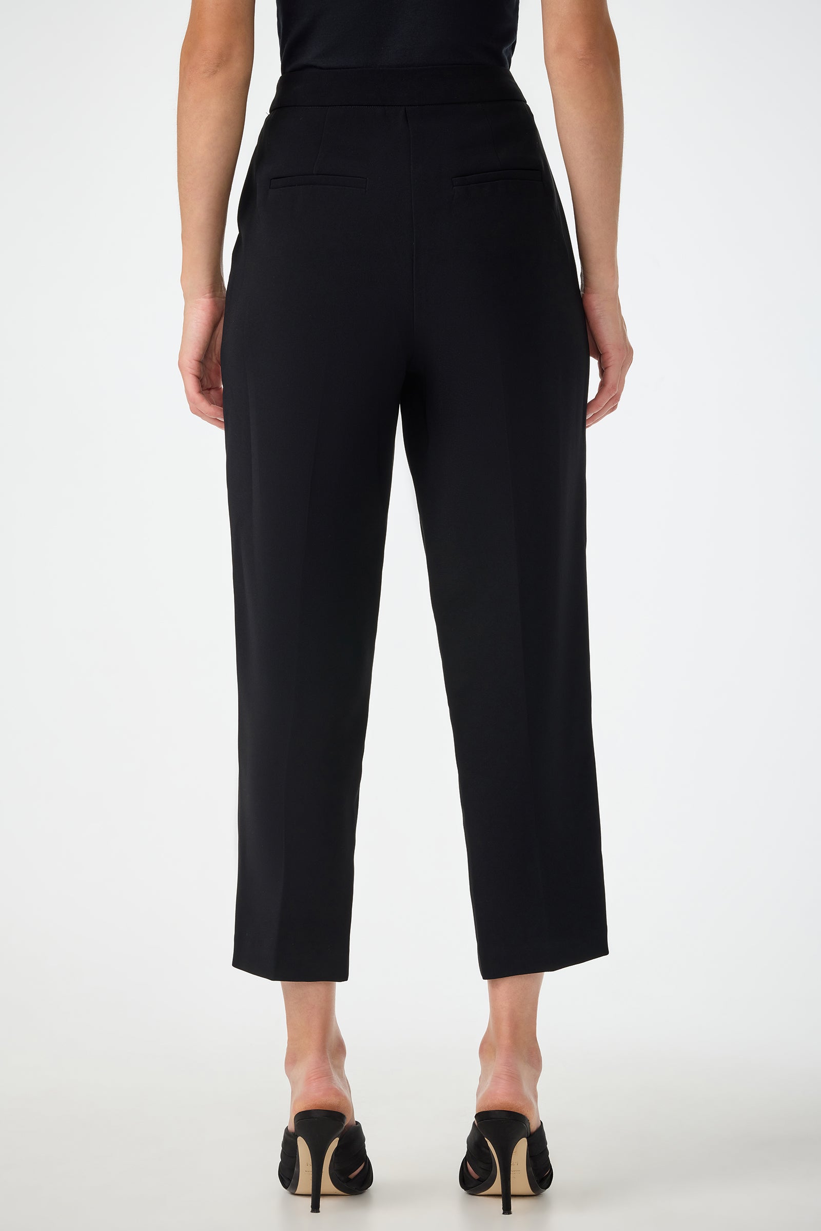 A person is seen from the back wearing the Larch Tailored Cropped Pants in black, featuring a high waist and two back pockets, paired with a black top and black heeled shoes against a plain white background.