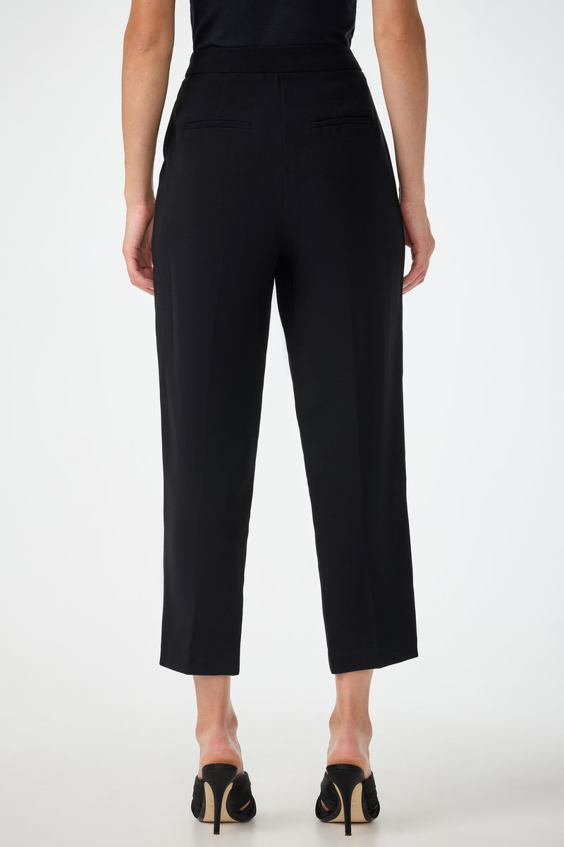 A person is seen from the back wearing the Larch Tailored Cropped Pants in black, featuring a high waist and two back pockets, paired with a black top and black heeled shoes against a plain white background.