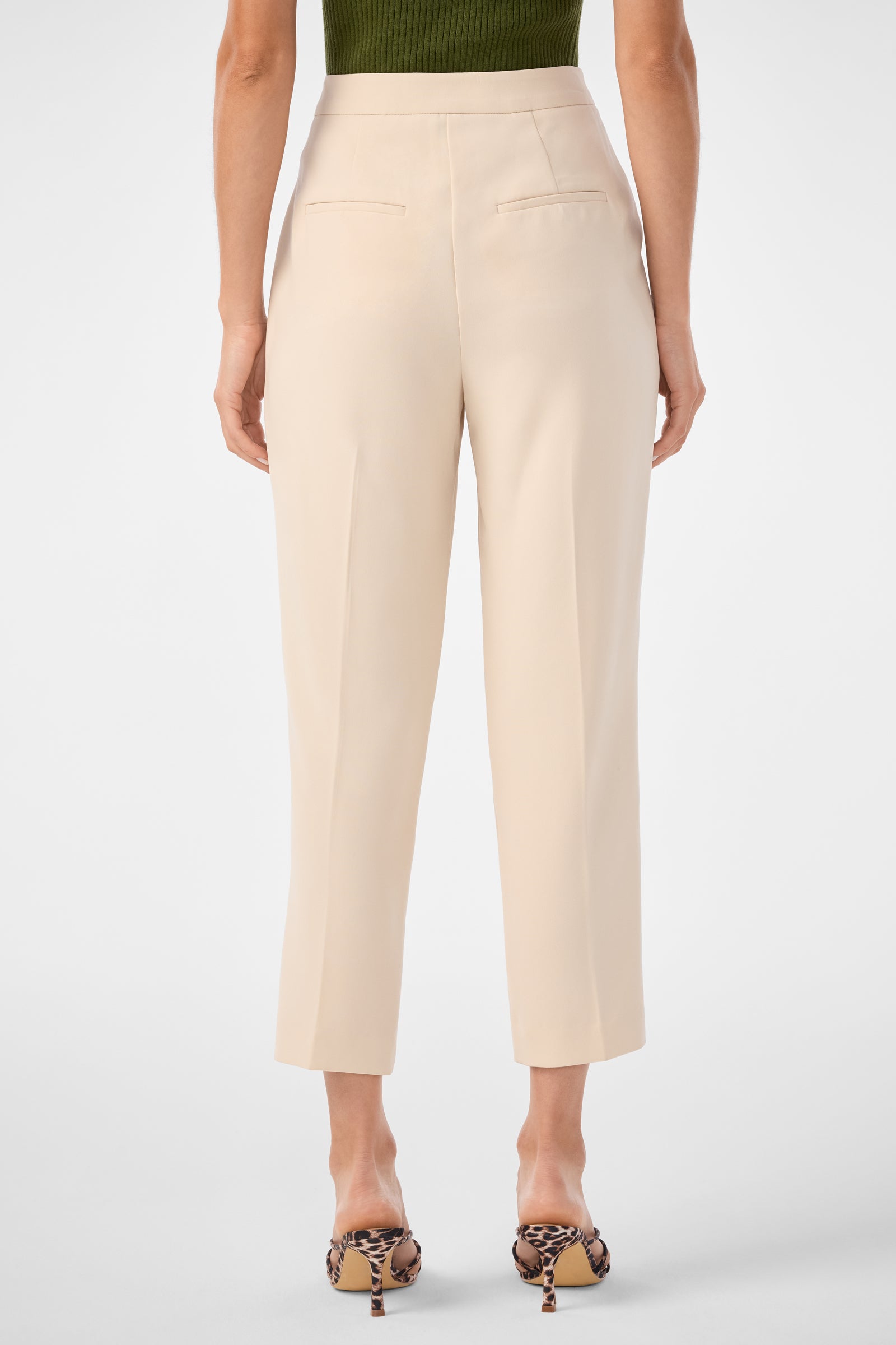 A person shown from the back wears Larch Tailored Cropped Pants in light beige, featuring high-waist, ankle length, and two back welt pockets, paired with leopard print heeled sandals and a dark green sleeveless knit top.