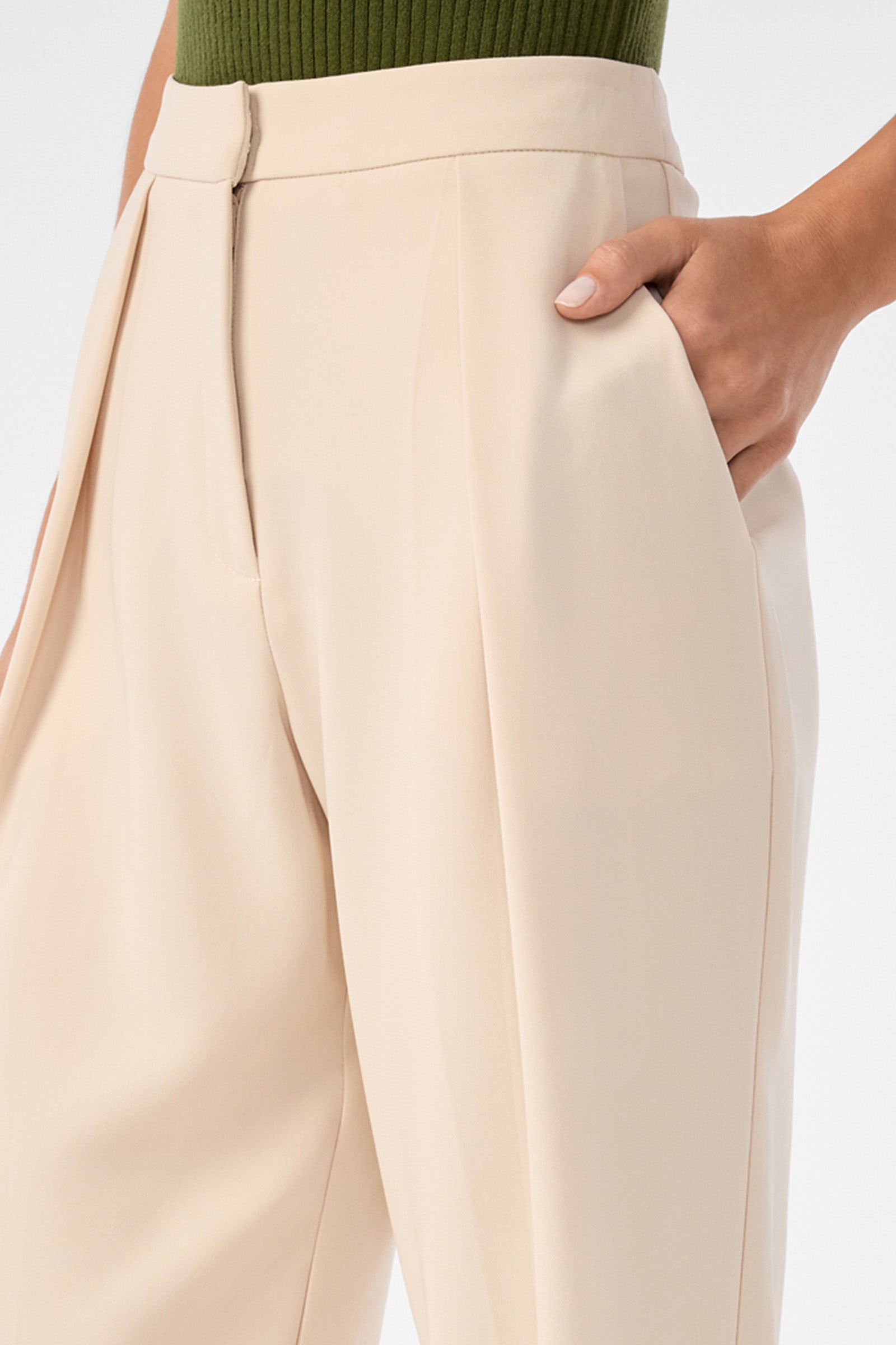 A person wearing the Larch Tailored Cropped Pants—high-waisted, beige tailored trousers with pleats—stands with one hand in their pocket, paired with a green ribbed top.
