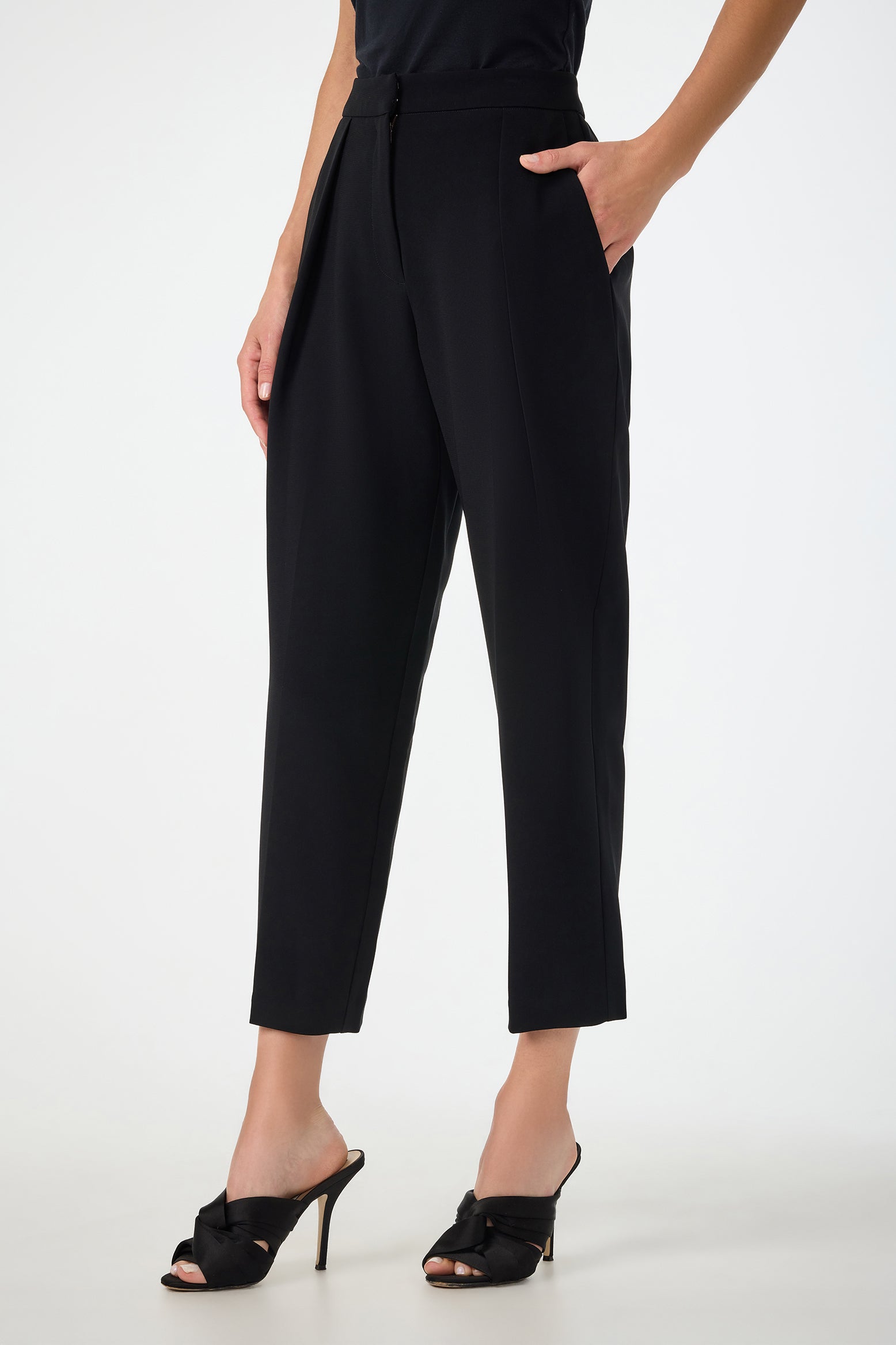 A person wears Larch Tailored Cropped Pants in black, high-waisted and tapered at the ankle, with hands in pockets, paired with black heeled sandals with bows, standing against a plain light background.