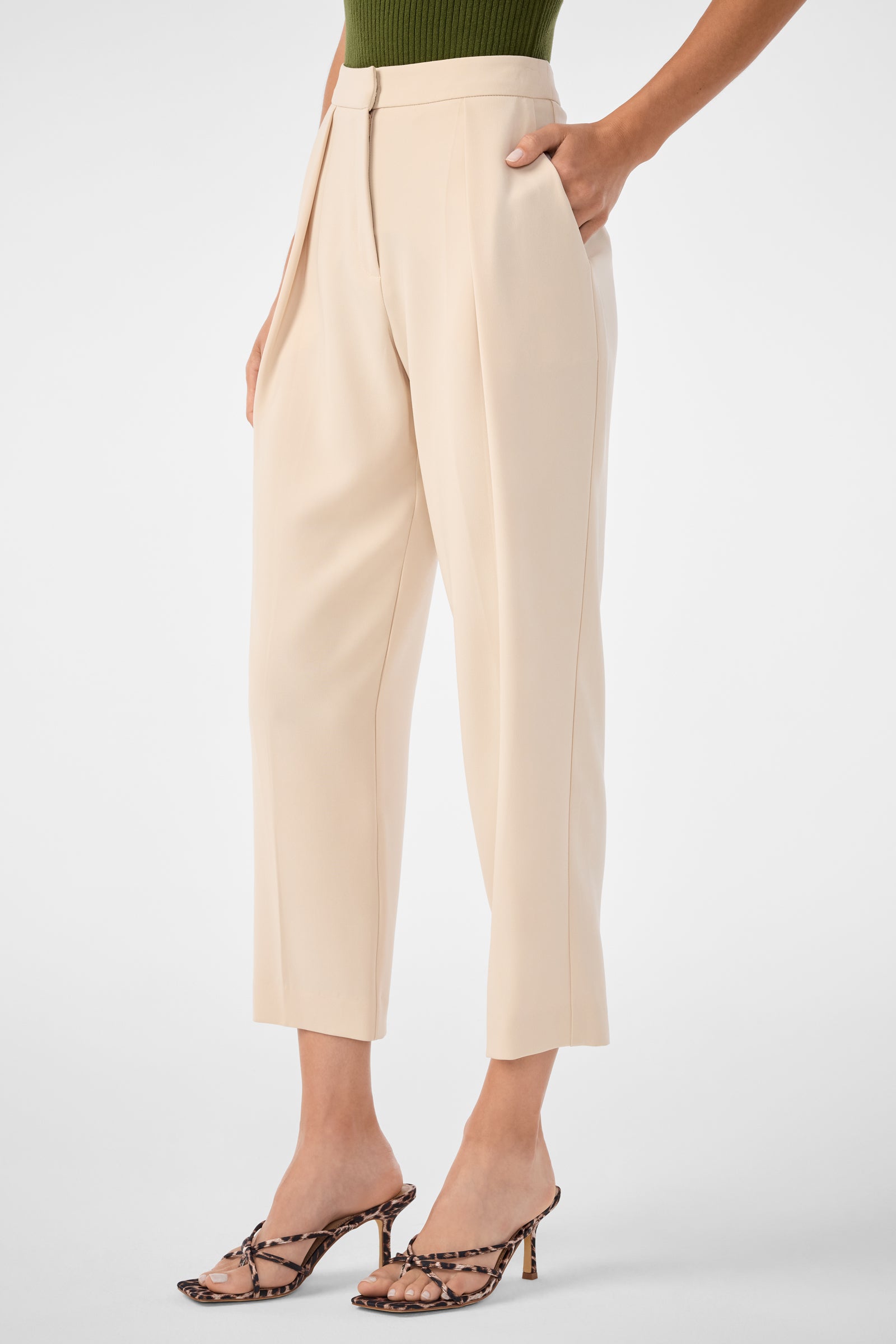 Wearing Larch Tailored Cropped Pants in beige, paired with a green top and snakeskin-patterned heeled sandals, the individual stands with one hand in their pocket against a plain background.