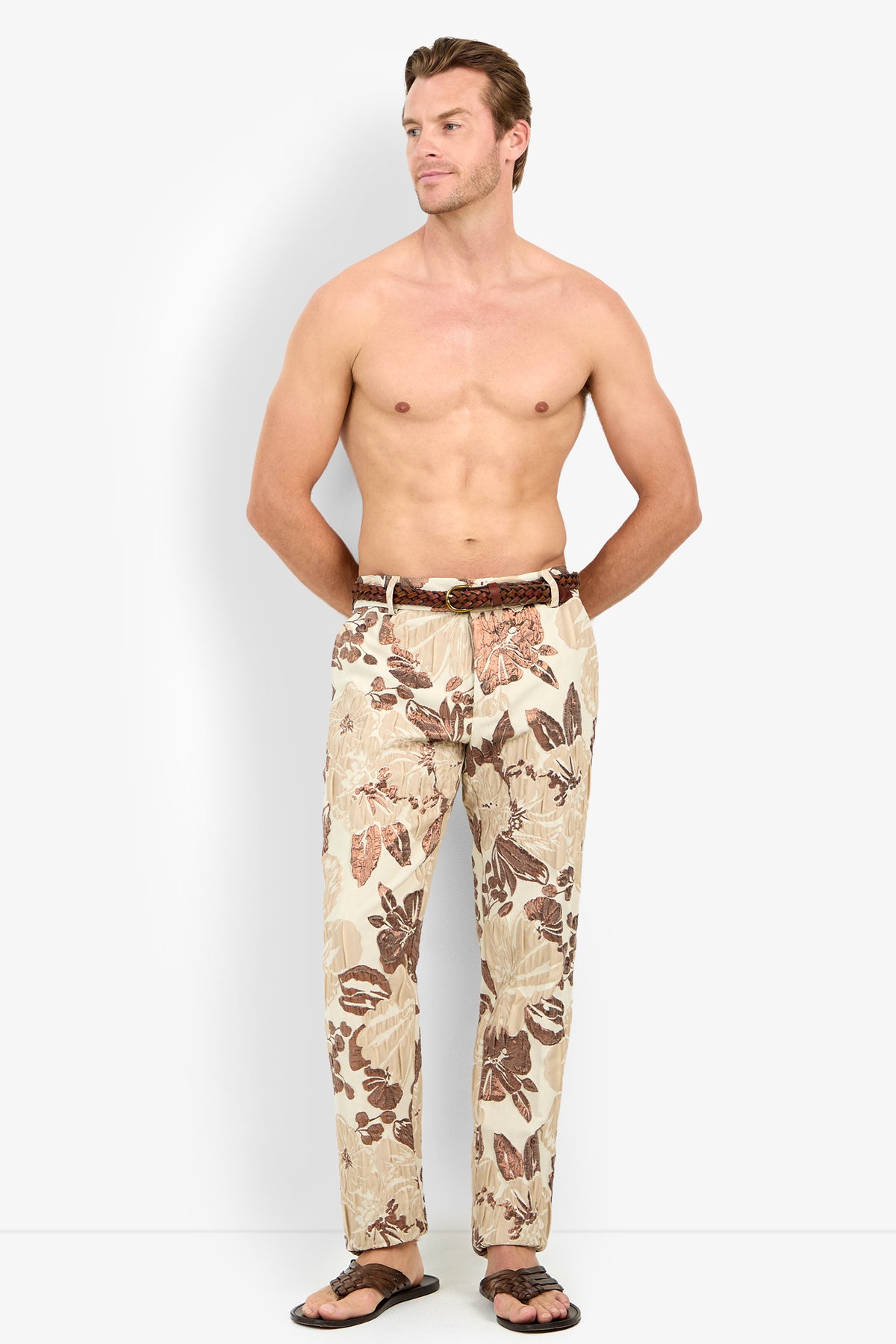 A shirtless man wears the Clyde Slim Trouser in beige floral print with a brown belt and dark sandals, standing against a plain white background, hands in pockets, looking slightly to the side.