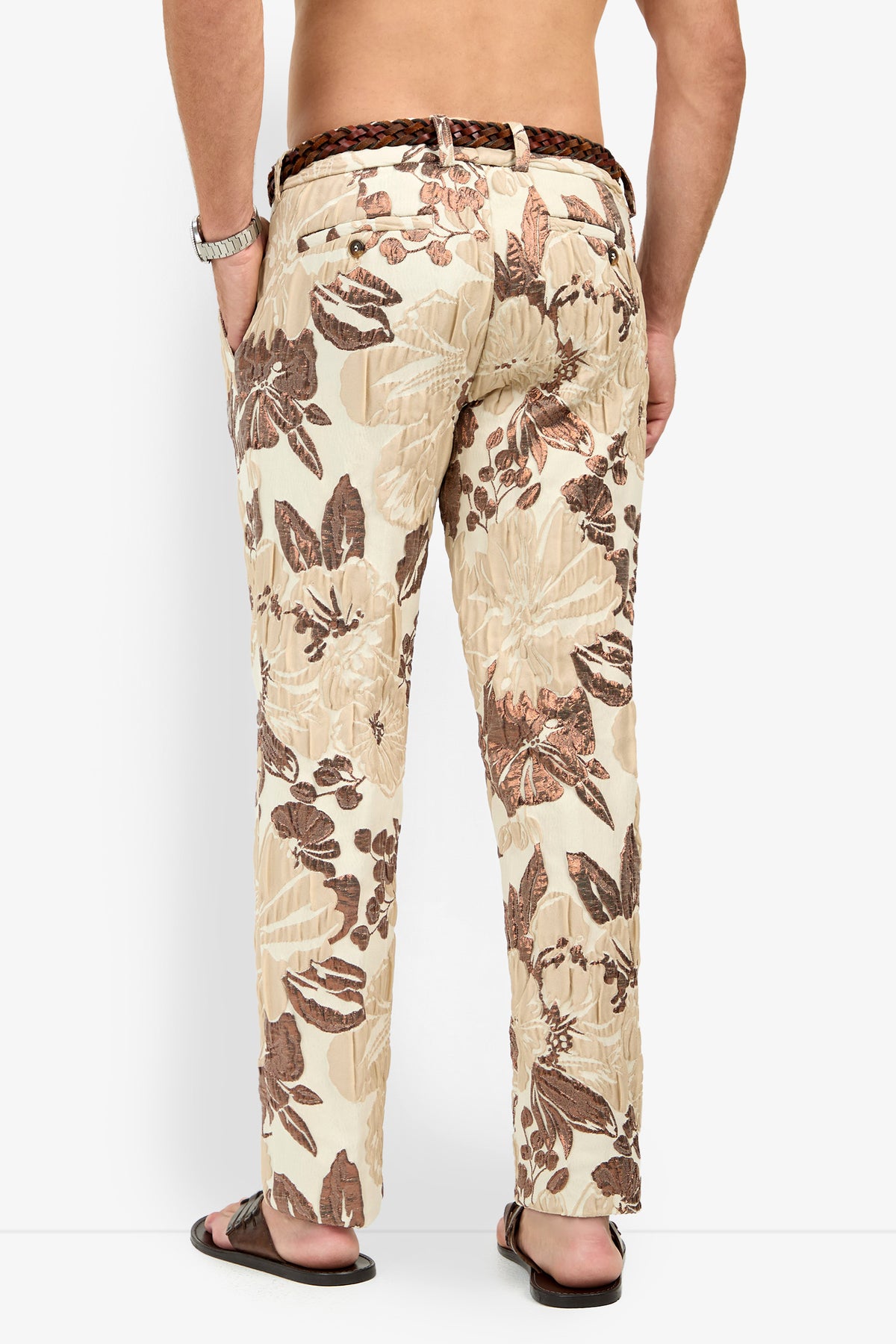 A man stands facing away, wearing Clyde Slim Trouser in cream with a brown floral pattern, paired with a brown braided belt, sandals, and a wristwatch. His hands are in his pockets.