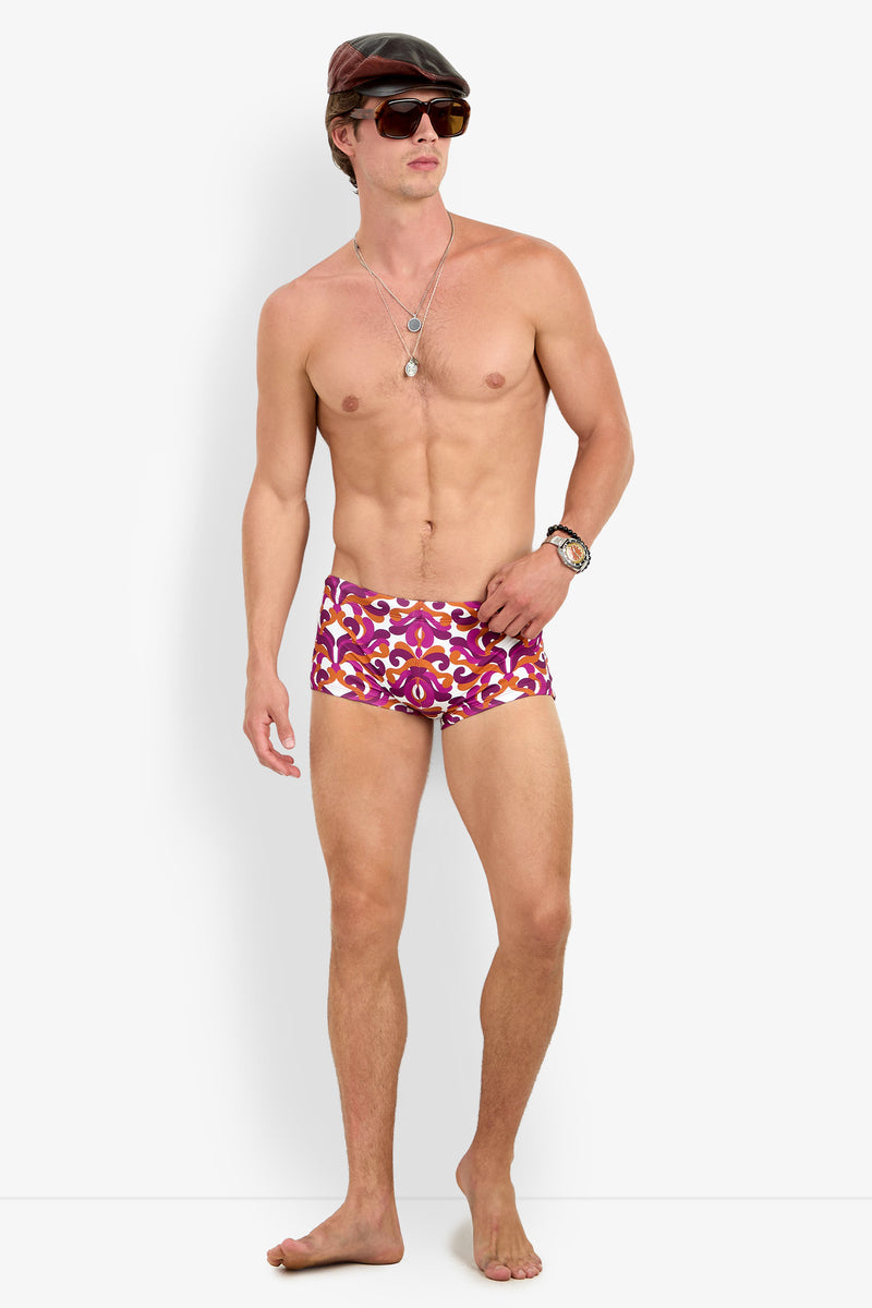 A shirtless man models the Jet Set Swim Trunk, paired with sunglasses, a cap, a wristwatch, and necklaces, standing barefoot against a plain white background.