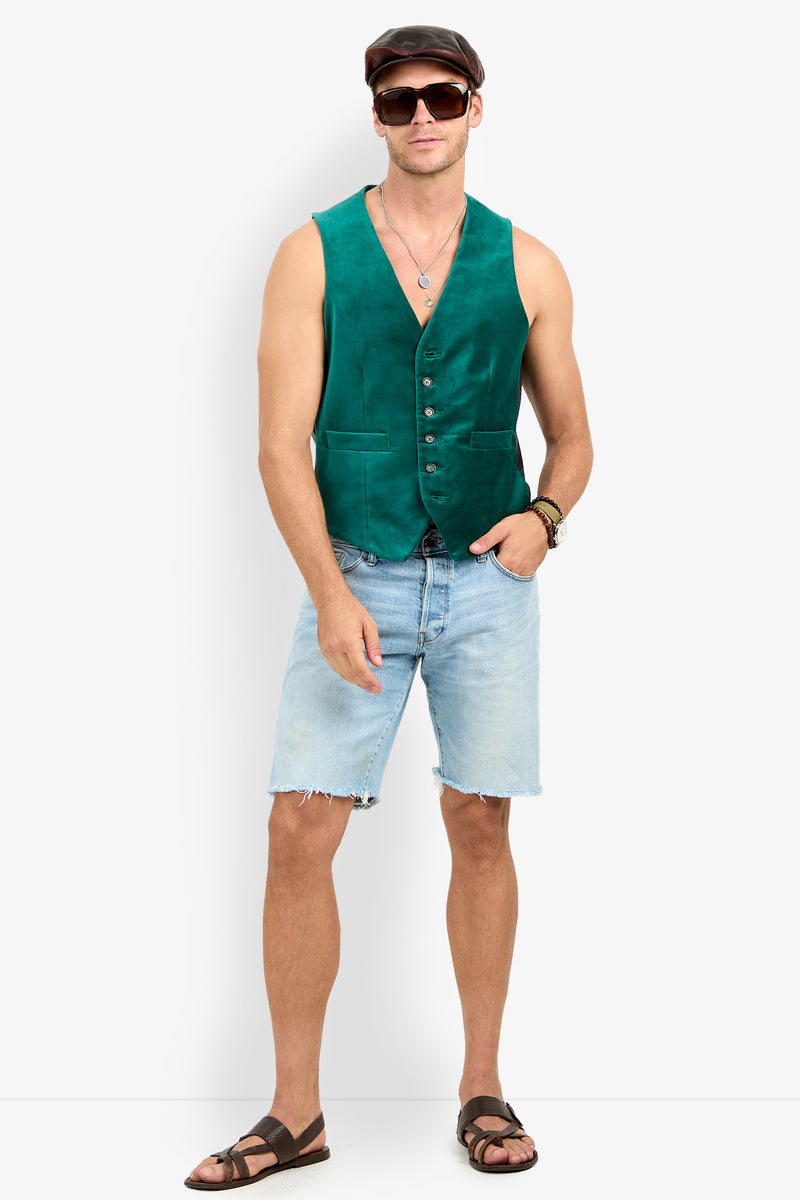 A man in an Irving Velvet Vest, light denim shorts, sandals, sunglasses, and a flat cap stands with one hand in his pocket against a white background.