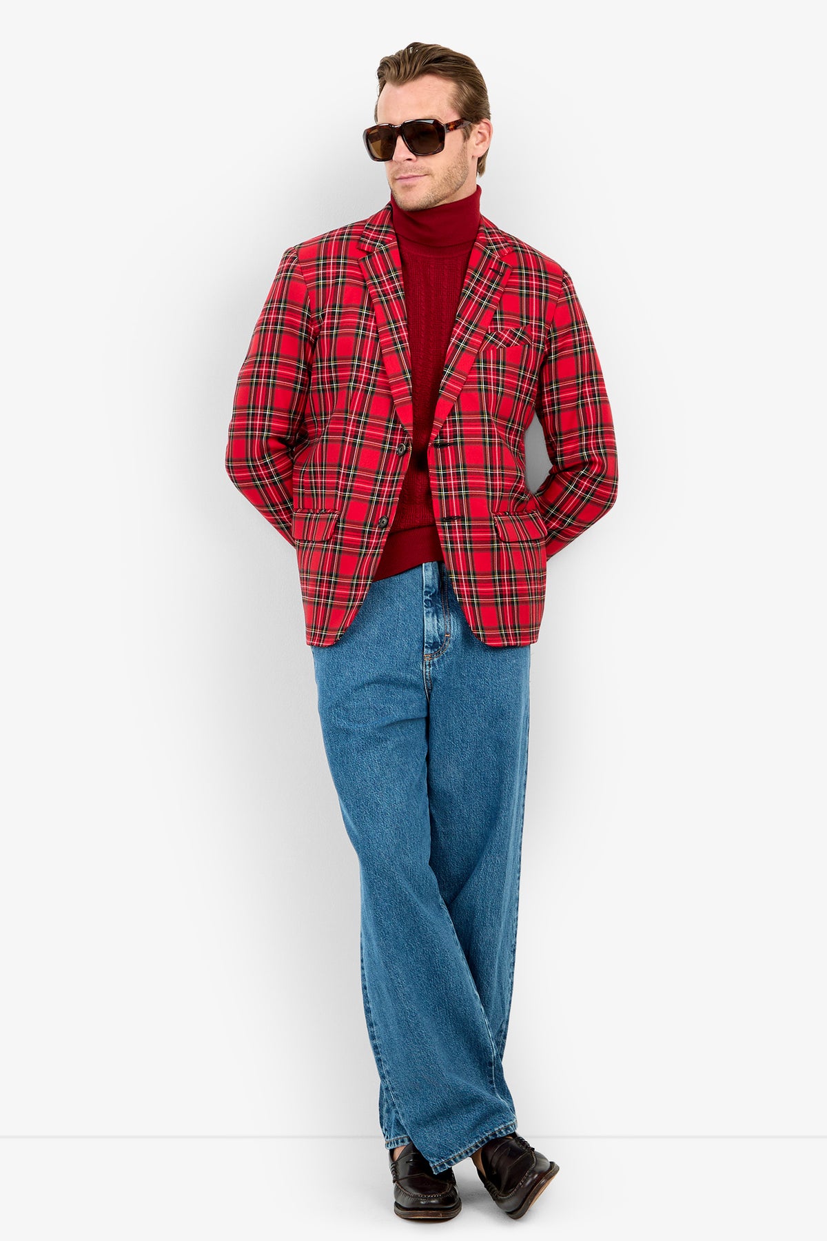 Man wearing sunglasses, the Thurston Blazer in red plaid, a red turtleneck, loose blue jeans, and black shoes stands relaxed and leans slightly while looking to the side against a plain white background.