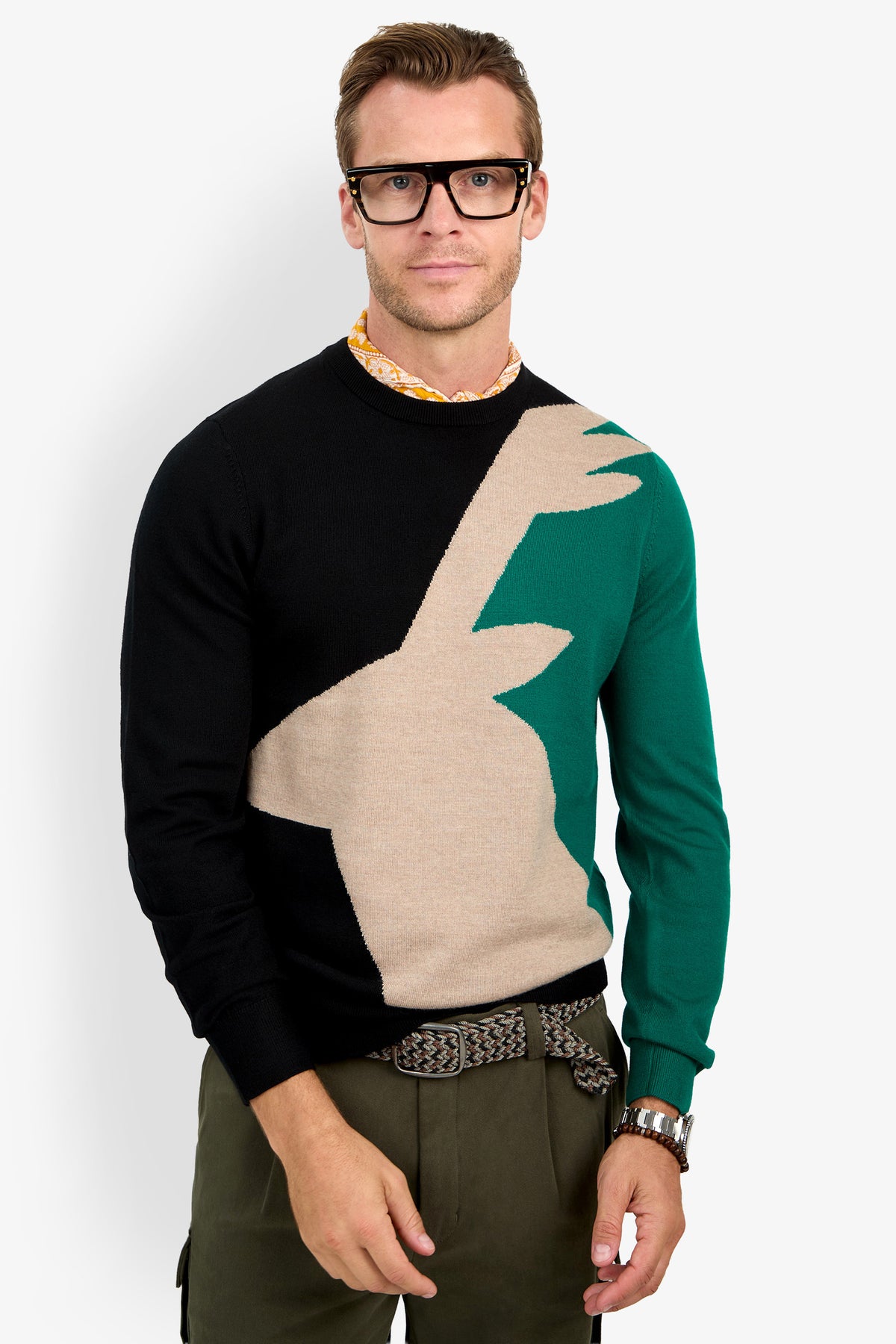 A man in thick black glasses, olive green pants, a patterned belt, and the Crewneck Sweater—featuring a multicolored abstract beige and green design—stands against a plain white background.