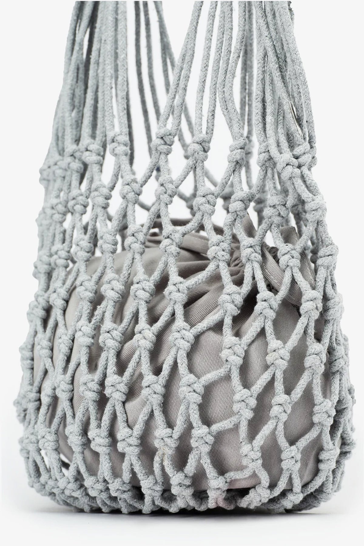 A close-up of the Nannacay Kim Woven Bag in light gray, featuring thick, knotted rope arranged in an open, diamond-shaped macramé pattern and holding a soft, light gray fabric pouch inside.