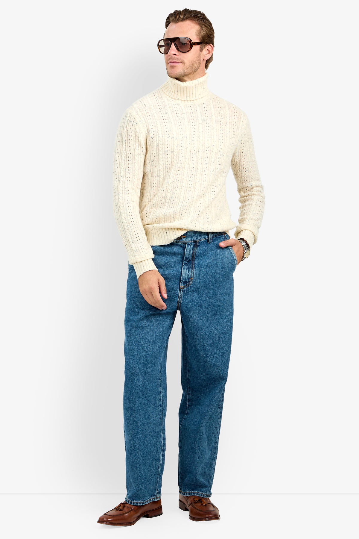 A man wearing large sunglasses, the Wesley Turtleneck Sweater, blue jeans, and brown shoes stands with one hand in his pocket against a plain background.