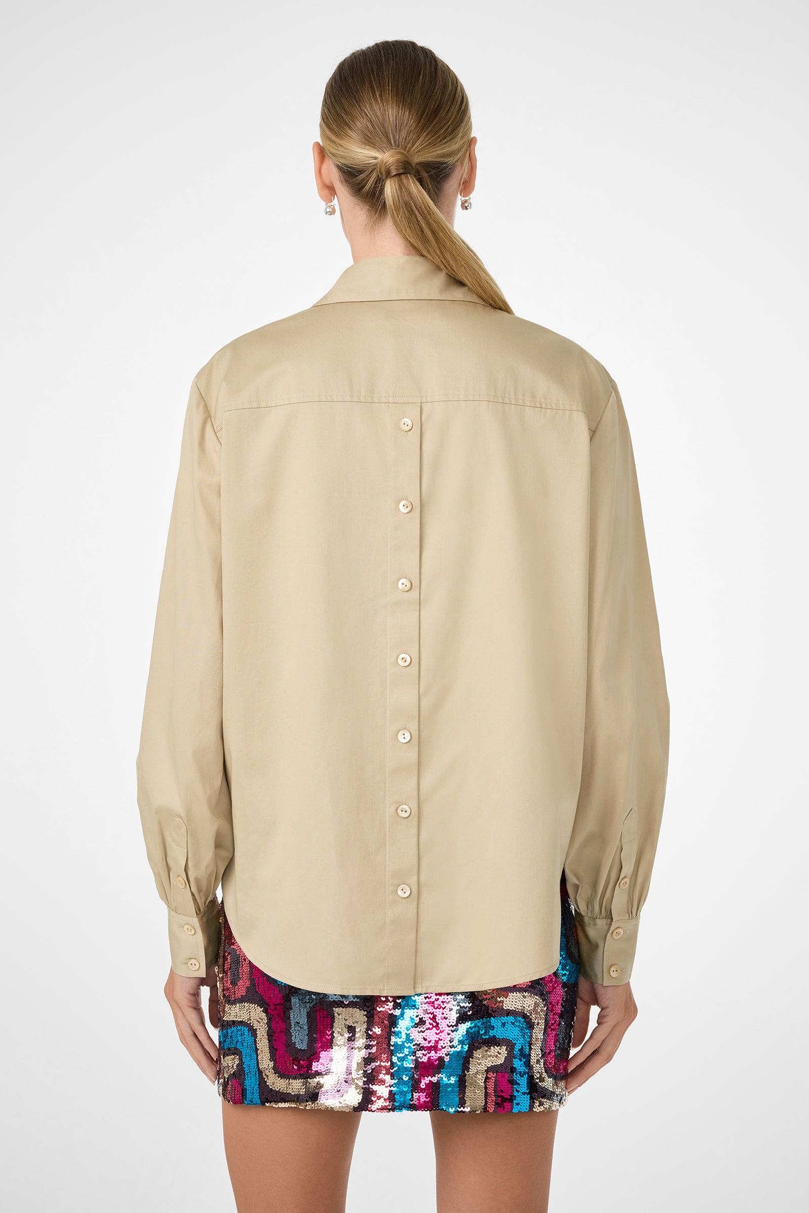 A woman stands facing away, wearing the Burle Top—a beige long-sleeve shirt with buttons down the back—and a colorful sequined geometric mini skirt. Her hair is tied back in a low ponytail.