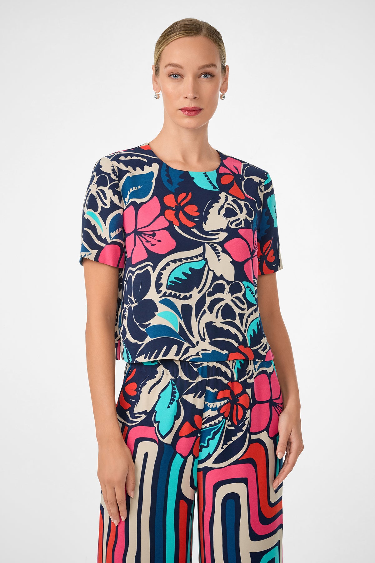 A woman wears the Sitio Top, a colorful short-sleeve shirt with bold floral patterns in navy, red, pink, and teal, paired with matching patterned pants. She stands against a plain light background and looks at the camera.