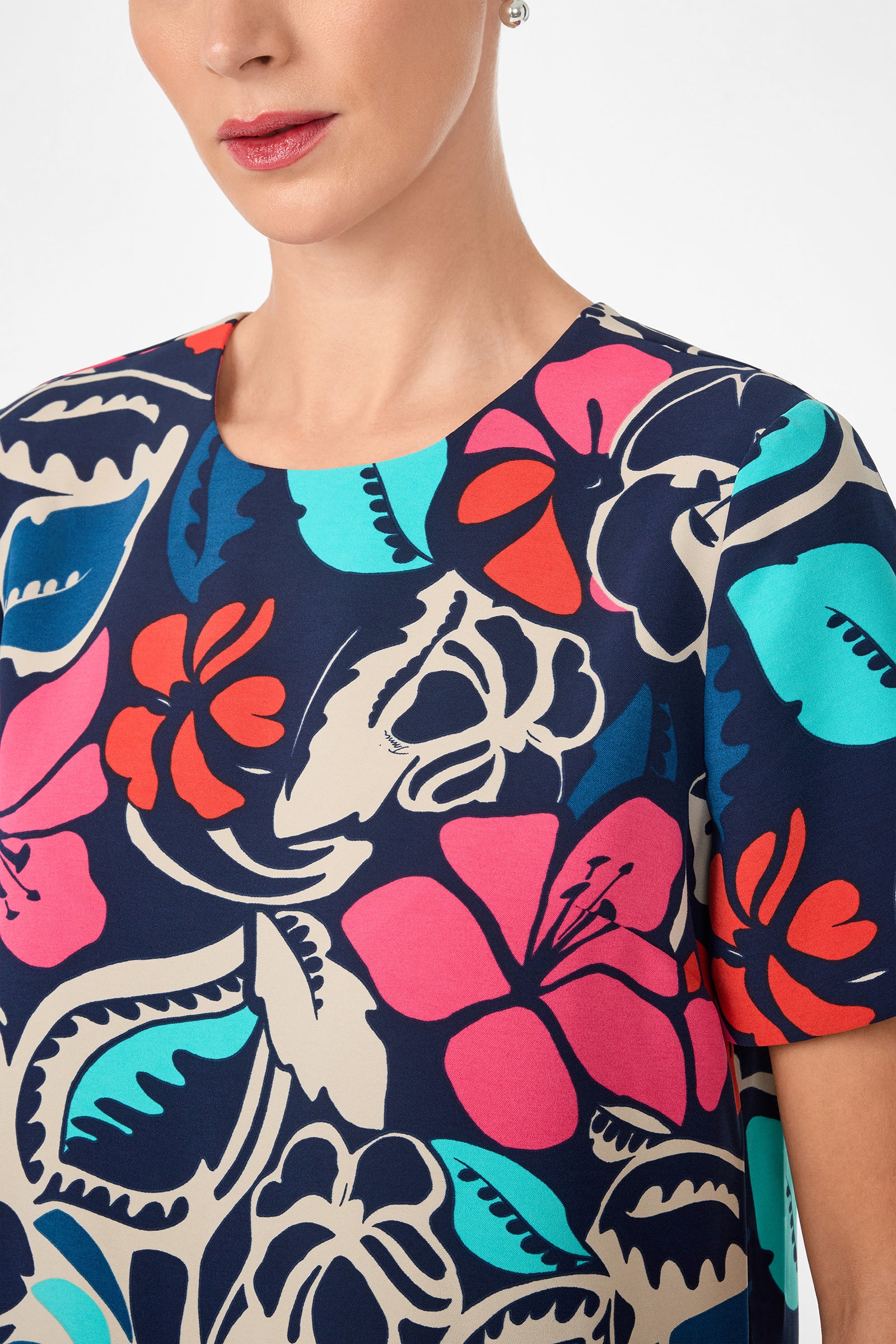 A woman wearing the Sitio Top, a short-sleeved style featuring a bold, colorful floral pattern in pink, blue, orange, and cream on a dark background. Her face is partially visible with pink lipstick and stud earrings.