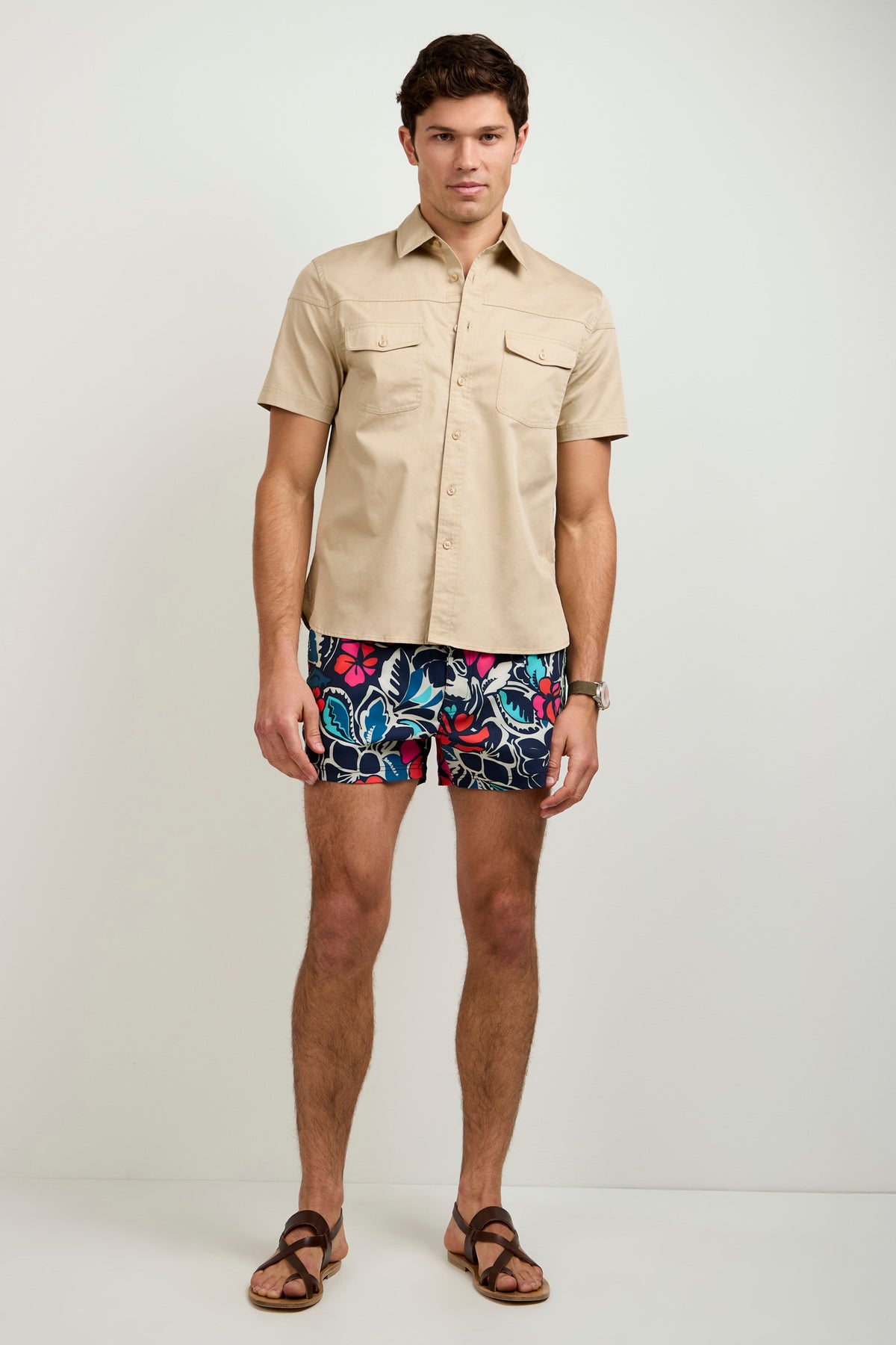 A man stands against a plain background wearing the Shelton Short-Sleeve Shirt in beige, paired with colorful floral shorts, brown sandals, and a watch.