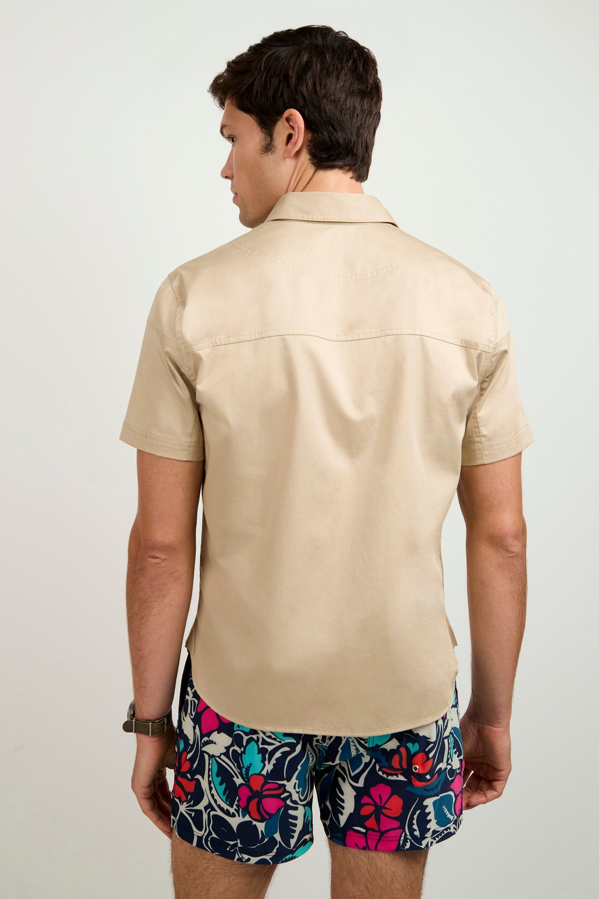 A man with short brown hair and a watch on his left wrist stands facing away, wearing the Shelton Short-Sleeve Shirt in beige and colorful floral shorts against a plain, light background.