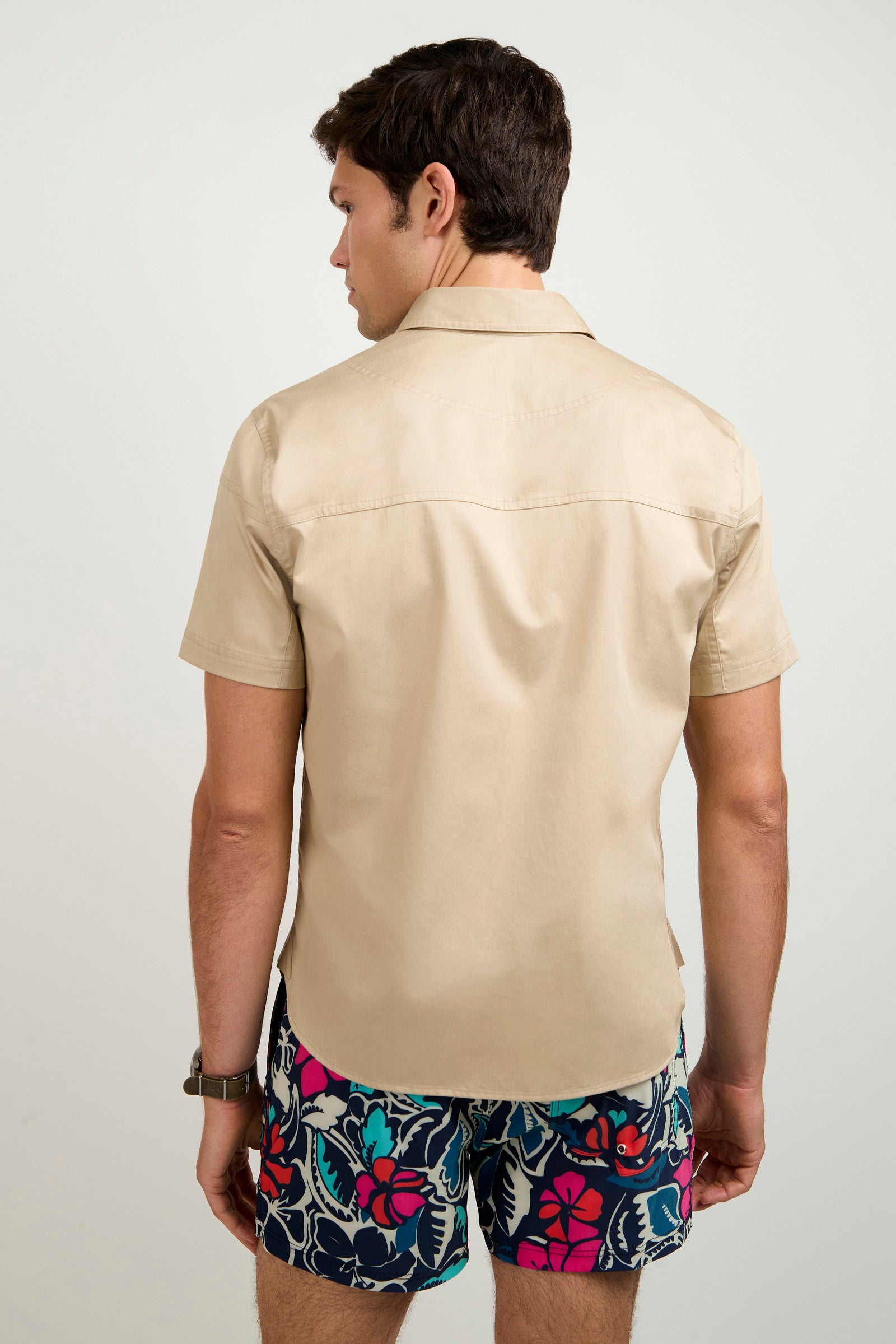 A man with short brown hair and a watch on his left wrist stands facing away, wearing the Shelton Short-Sleeve Shirt in beige and colorful floral shorts against a plain, light background.