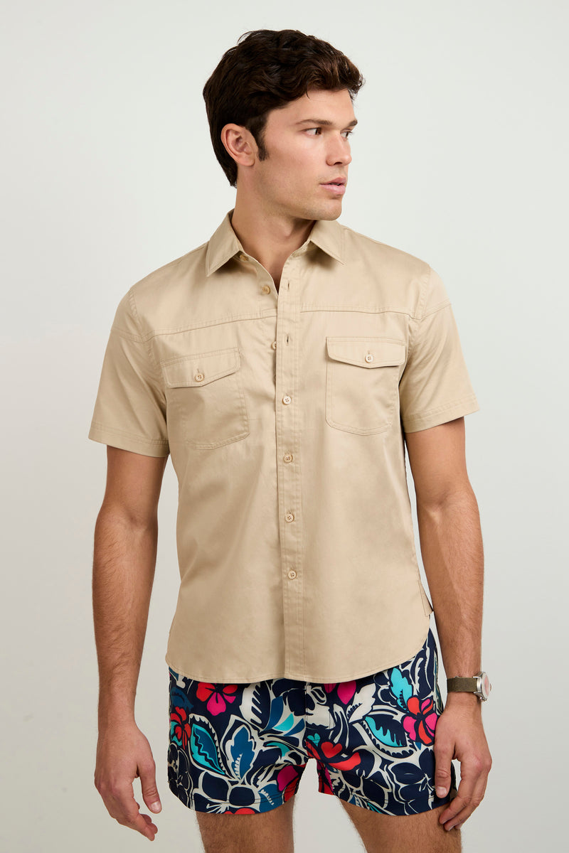 A man in the Shelton Short-Sleeve Shirt stands against a plain light background, looking slightly to his left. He pairs the beige button-up with breast pockets and colorful floral swim trunks.