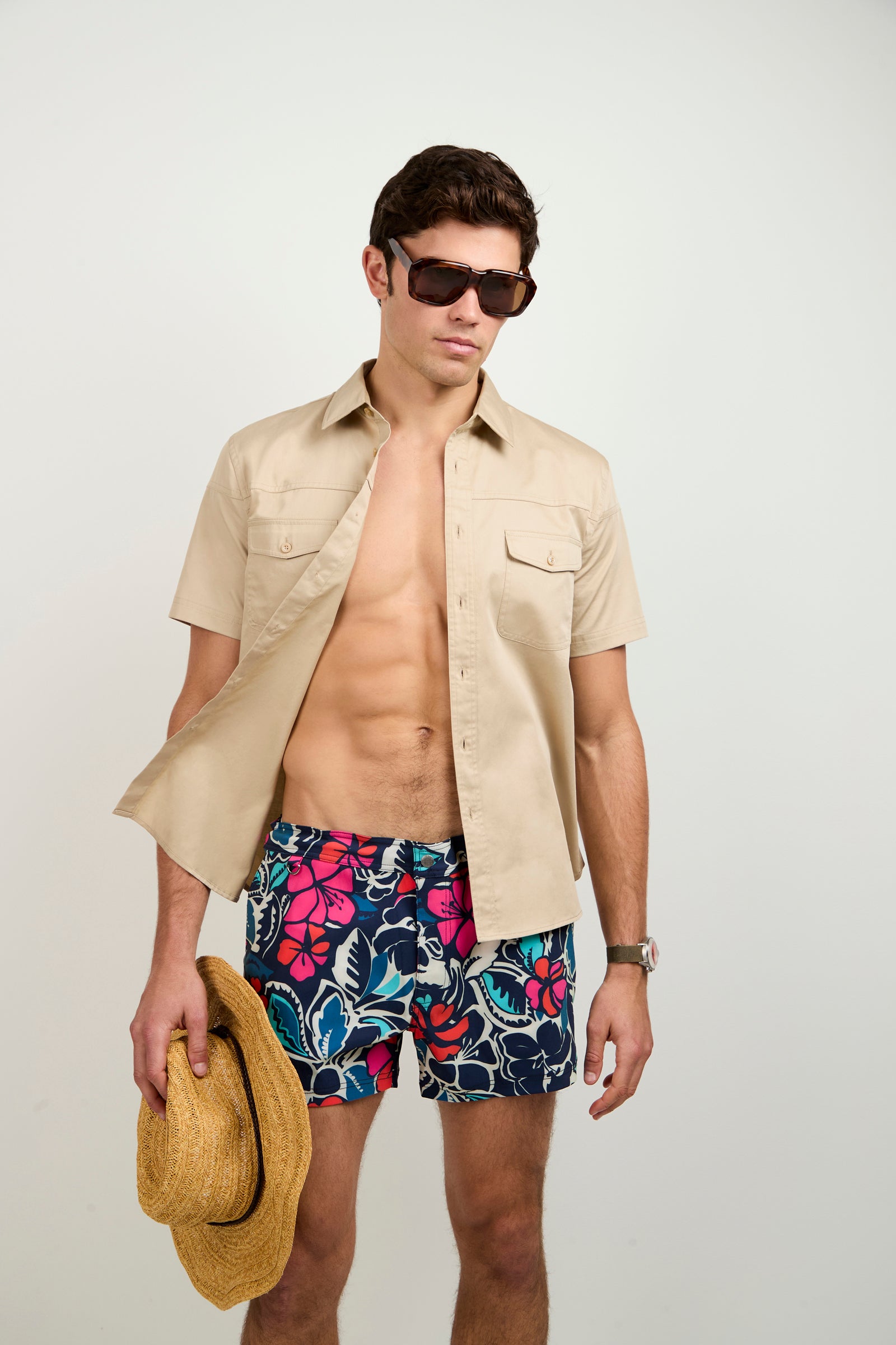A man models the Chico Swim Trunk - Copacabana, paired with a beige short-sleeve button-up shirt and sunglasses, holding a straw hat at his side against a plain white background.