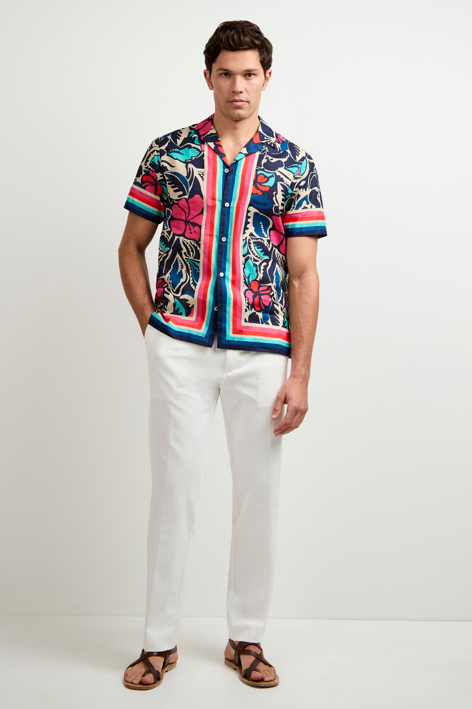 A man with short dark hair stands against a plain light background, wearing a colorful floral button-up, the Clyde Slim Trouser - Stretch Piqué in white, and brown sandals. He has his hands in his pockets and a neutral expression.