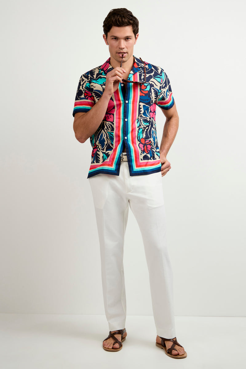 A man stands against a plain background wearing the Josue Short-Sleeve Shirt - Copacabana with white pants and brown sandals, holding sunglasses near his mouth and looking forward with a relaxed expression.