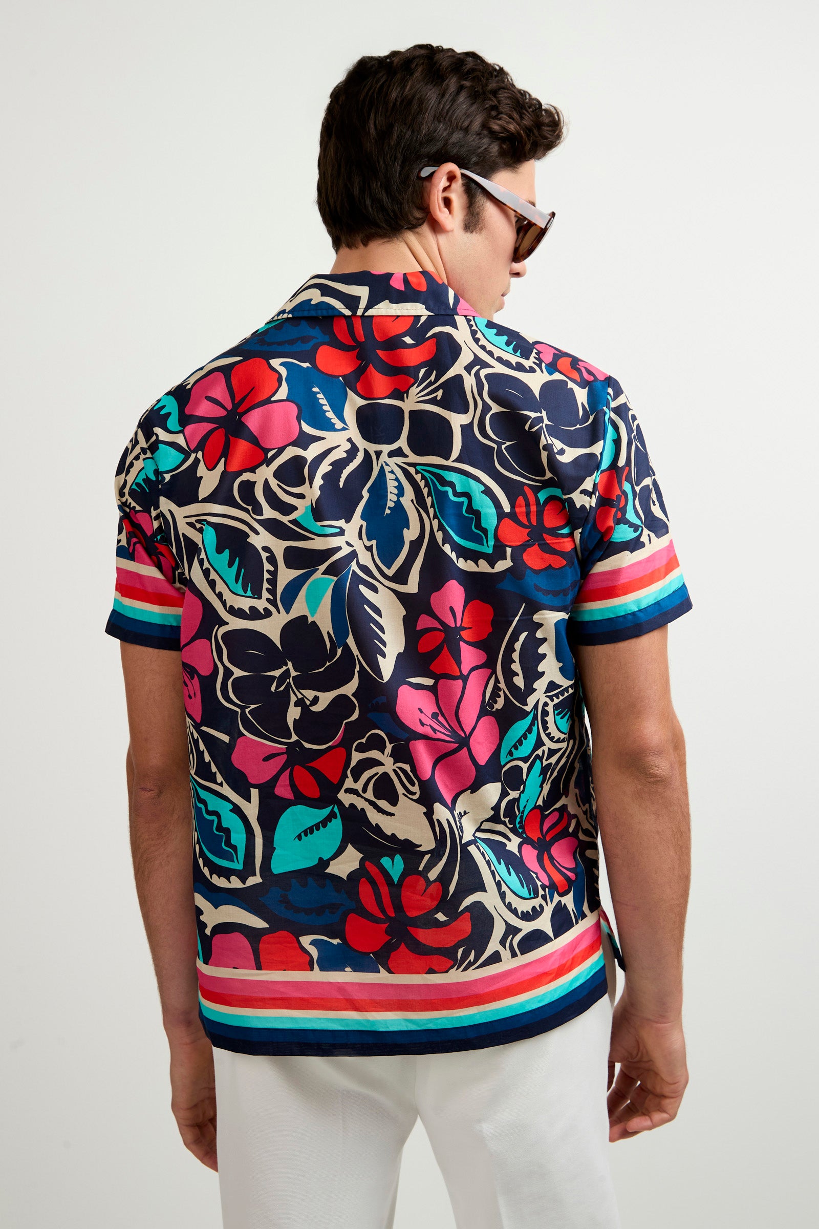 A man with short dark hair and sunglasses, seen from behind, wears the Josue Short-Sleeve Shirt - Copacabana featuring a bold floral pattern with stripes, paired with white pants.