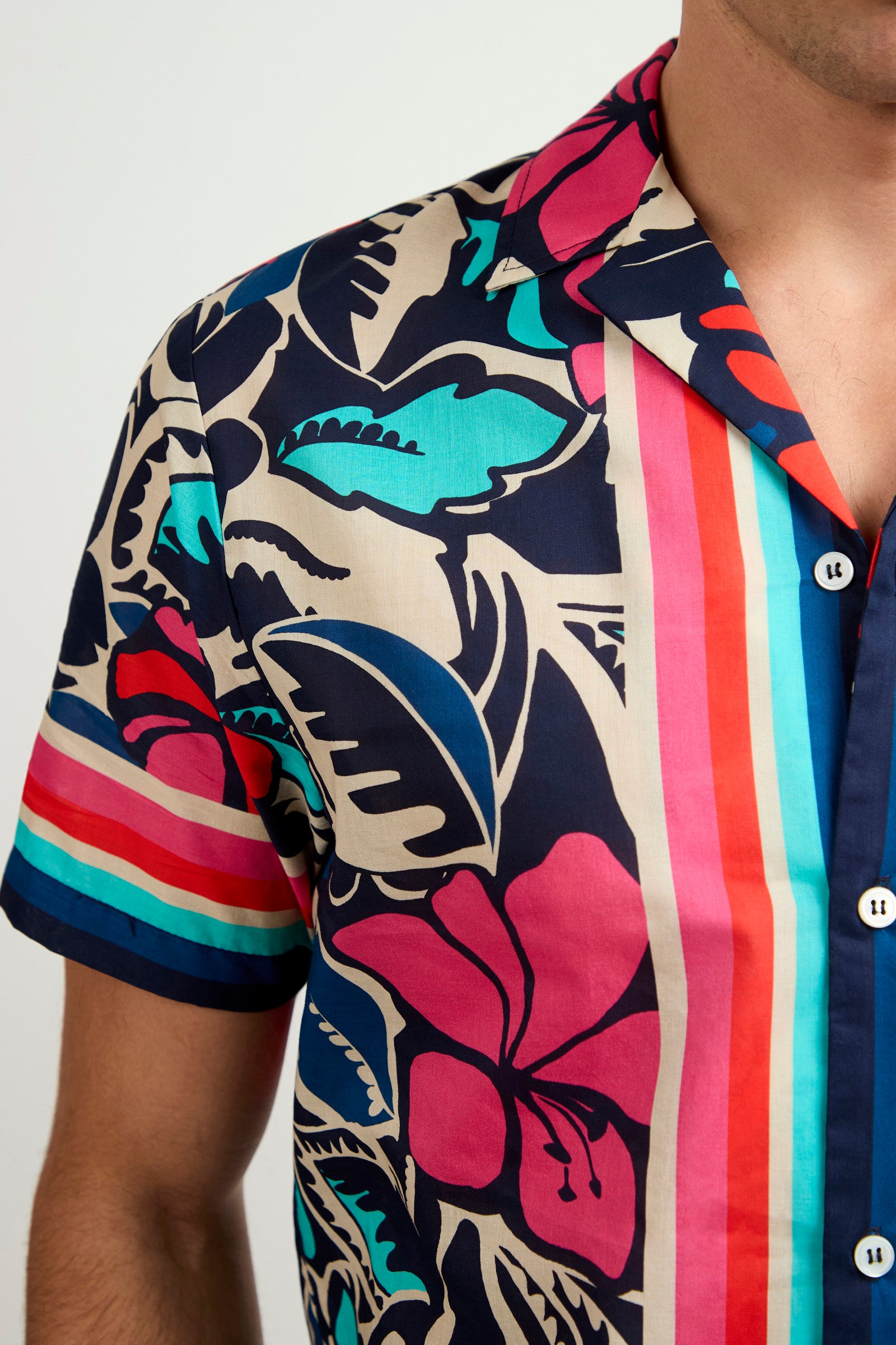 Someone wearing the Josue Short-Sleeve Shirt - Copacabana, which has a vibrant tropical floral design with bold leaves, flowers, and vertical blue, red, and beige stripes. Only the upper torso is visible.