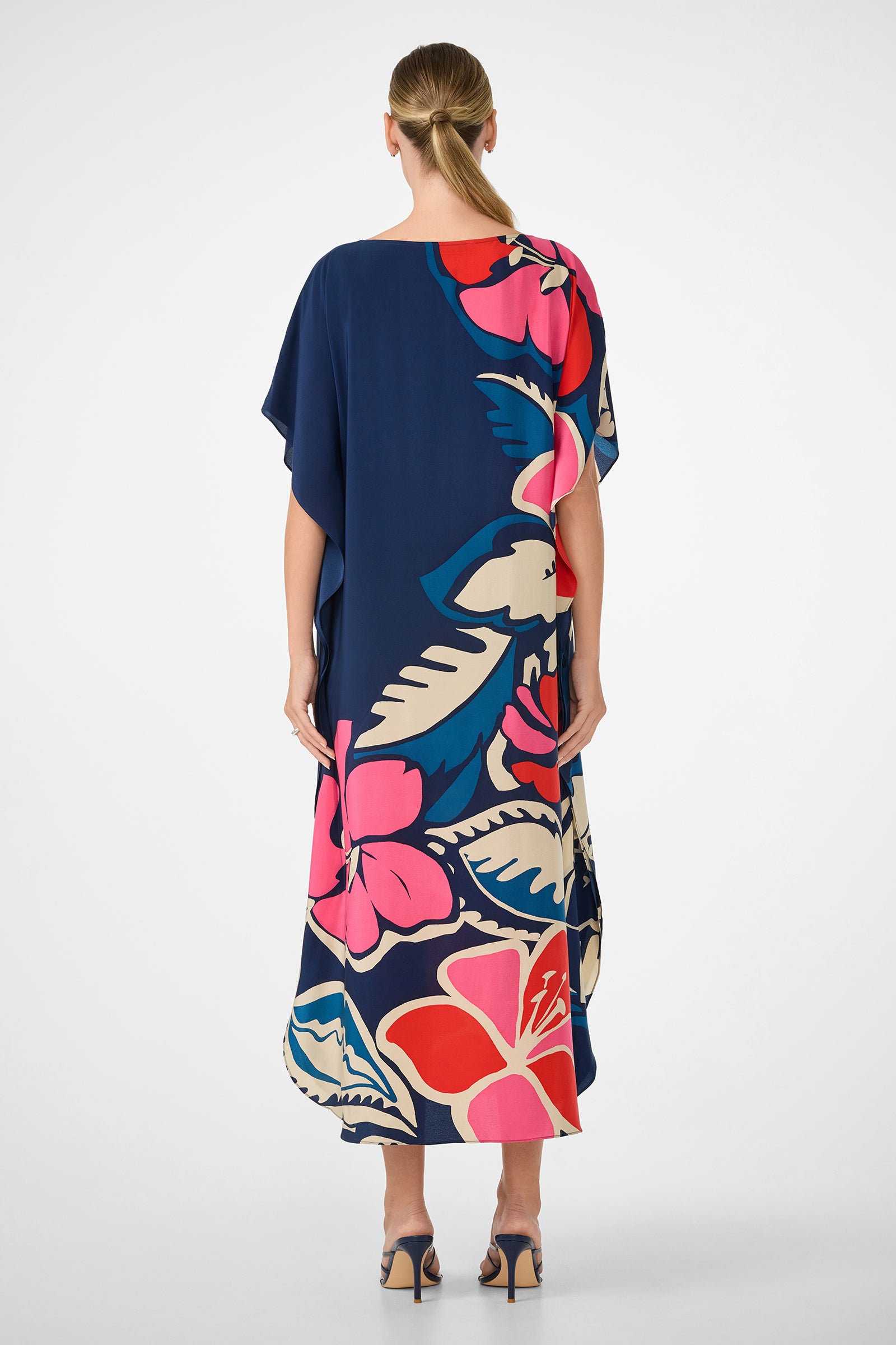 A woman with blonde hair in a low ponytail faces away, wearing the Jalani Maxi Caftan Dress—an ankle-length, navy blue dress featuring oversized pink, red, white, and blue floral prints. She pairs it with high-heeled sandals.