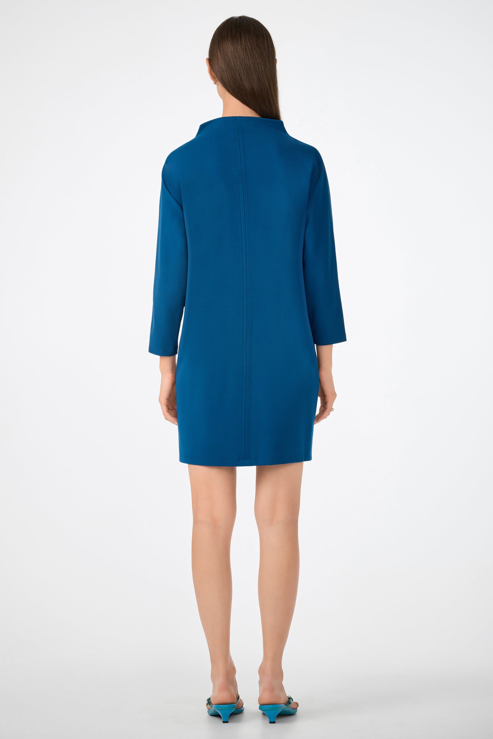 A woman with straight brown hair stands facing away in the Caracas Dress— a knee-length, long-sleeved teal dress— and matching teal pointed-toe heels against a plain white background.