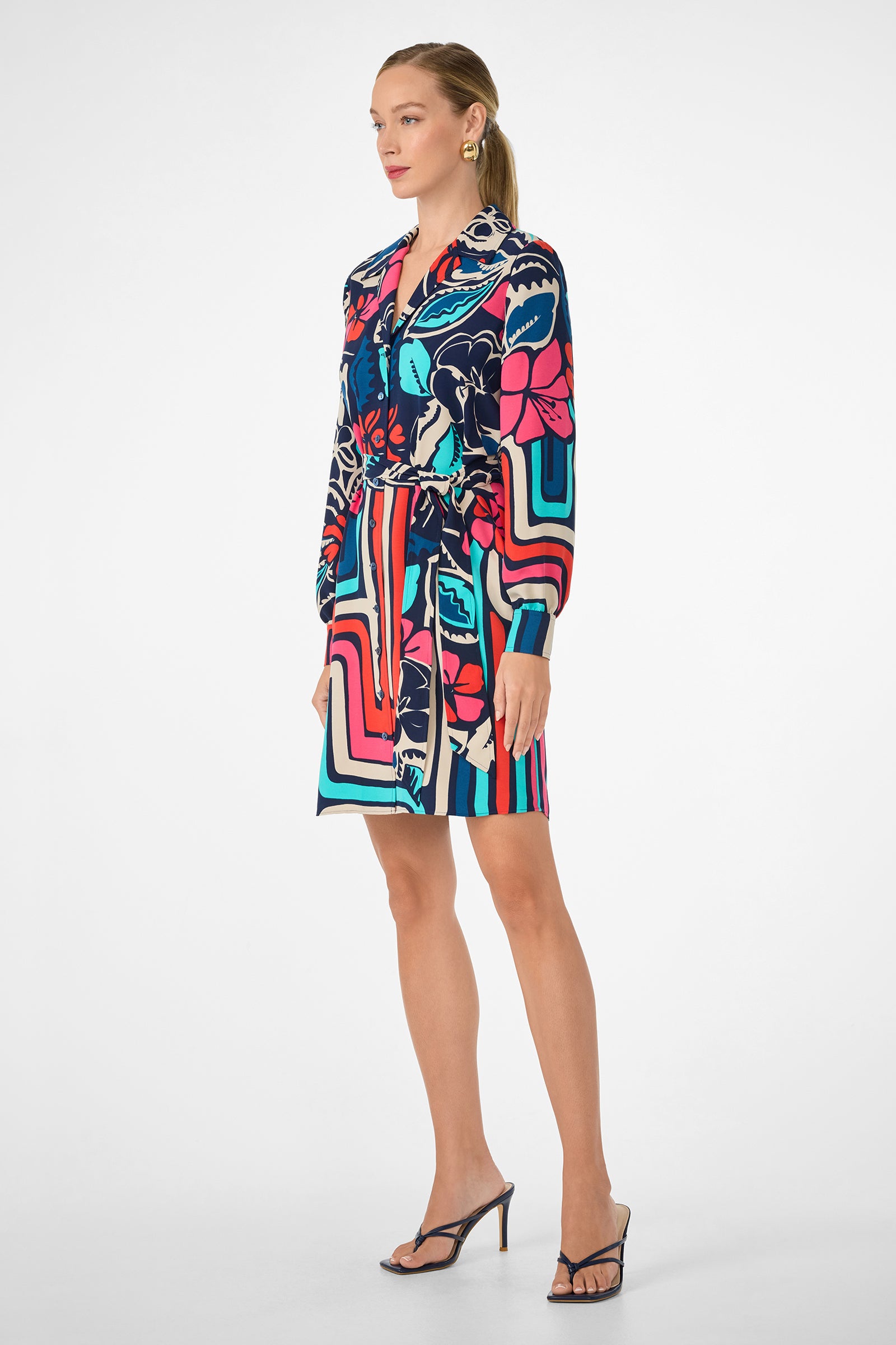 A woman poses against a white background in the Flamenco Dress, a colorful patterned long-sleeve style with abstract floral and geometric designs in blue, pink, red, and cream. She pairs it with black heeled sandals and gold earrings.