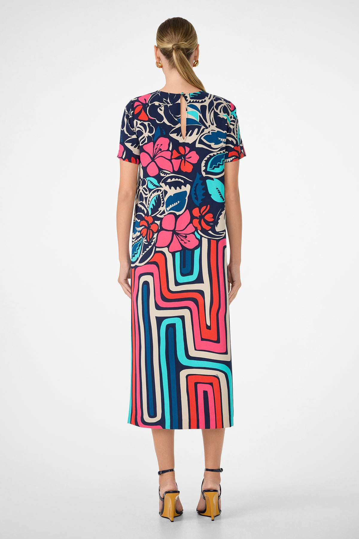 A woman with blonde hair in a low bun wears the Martay Dress, featuring a bold floral top and geometric striped skirt. She pairs it with strappy heels and stands against a plain white background, facing away from the camera.