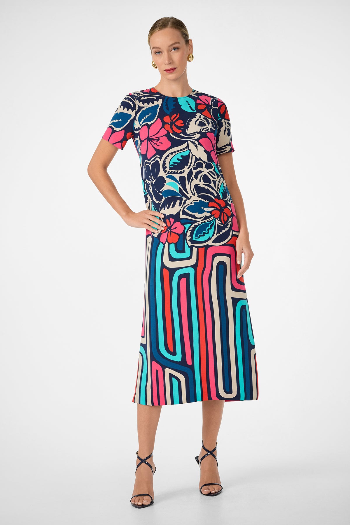 A woman models the Martay Dress, a colorful short-sleeve midi with bold red, blue, pink, and cream floral and geometric patterns. She pairs it with black strappy heels and gold earrings against a plain white background.