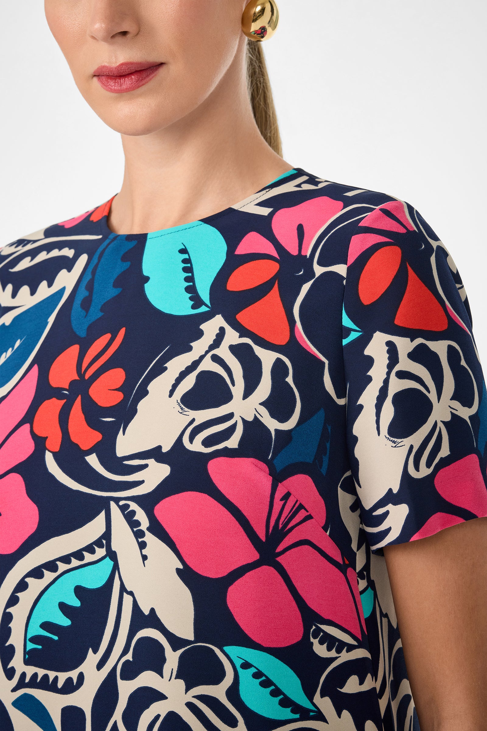 A woman wears the Martay Dress, a navy blue top with a vibrant floral print in red, pink, blue, beige, and teal. She has red lipstick and gold earrings. The image captures her from the shoulders up against a plain background.