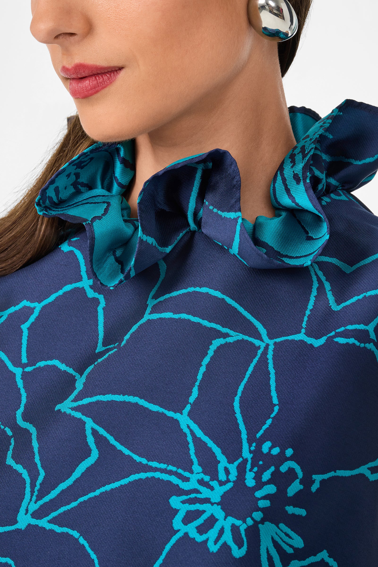 A woman wearing bold silver earrings and the Edina Dress, a navy blue top with teal floral patterns and a ruffled collar. Only her lower face and upper torso are visible.